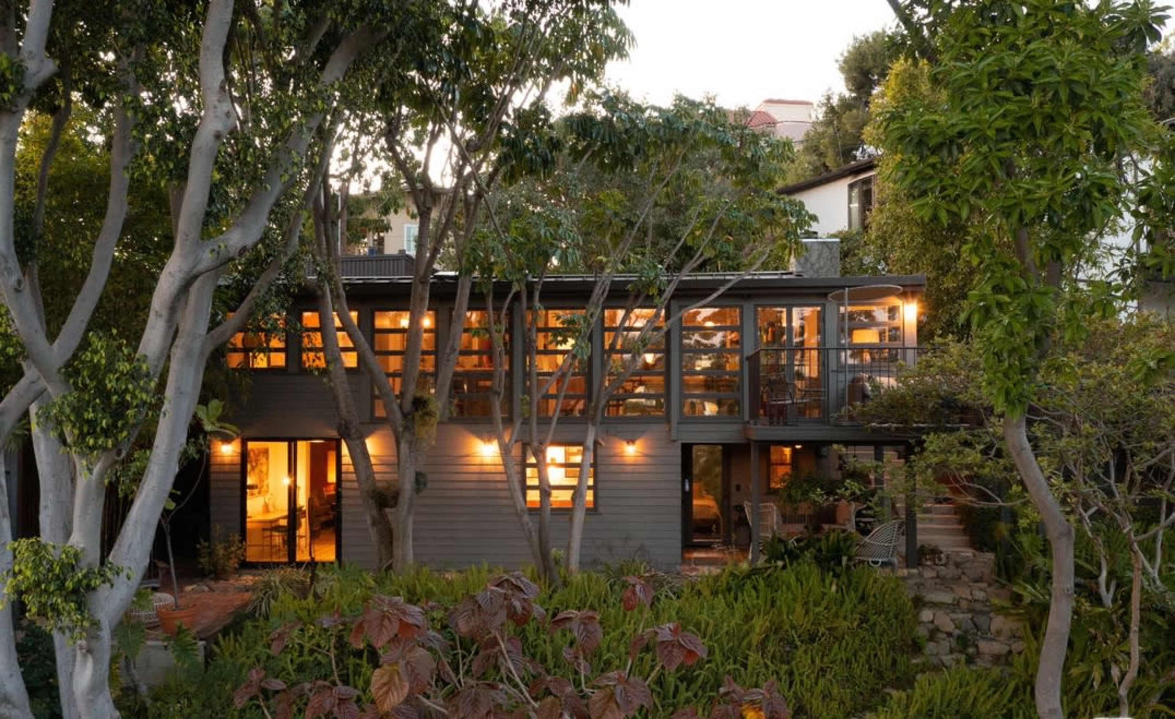 A modern two-story house is surrounded by trees and gardens, with warm lights glowing from its large windows at dusk.