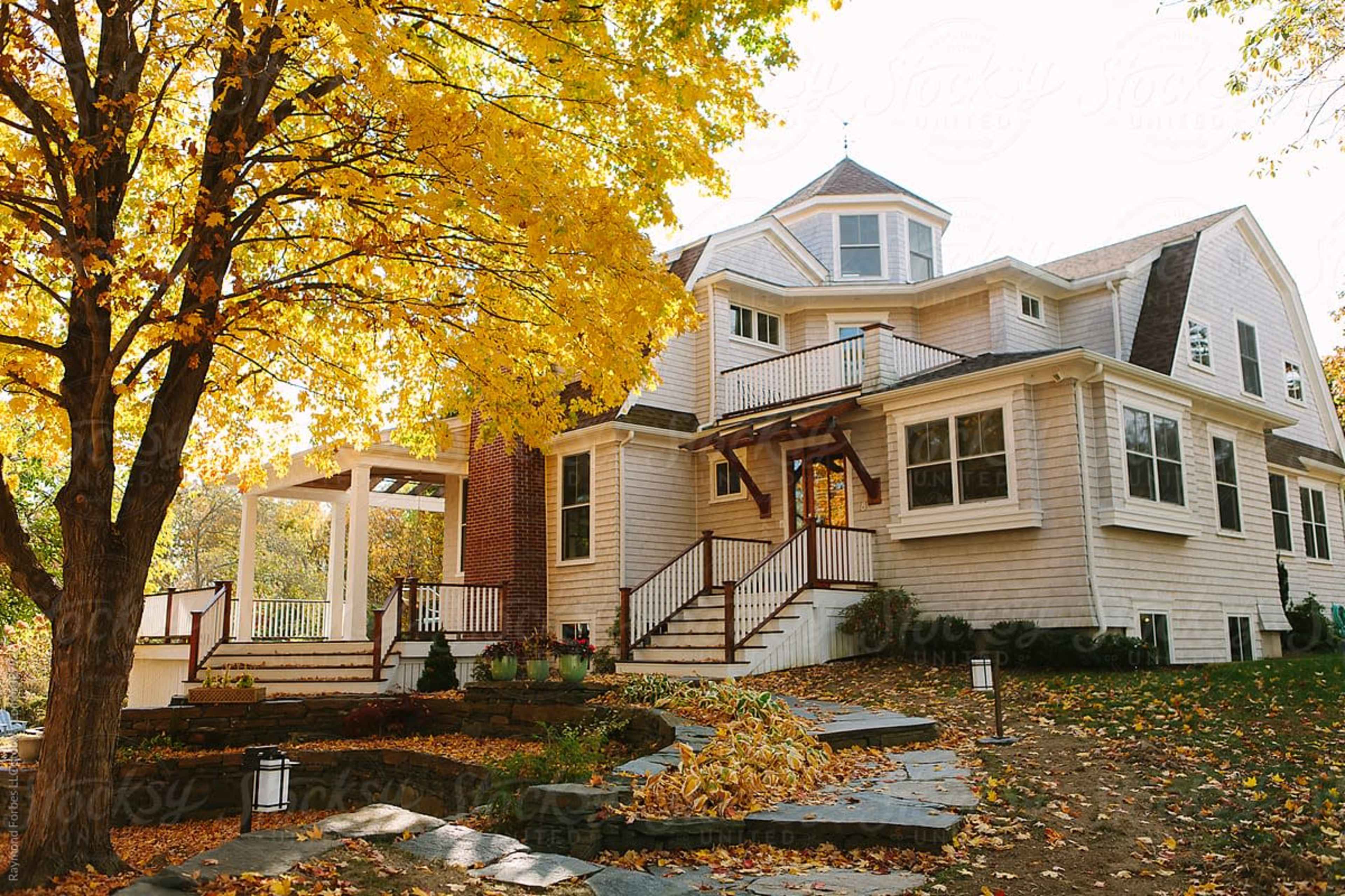 A two-story house with a light-colored facade and a porch is surrounded by trees displaying vibrant autumn foliage.