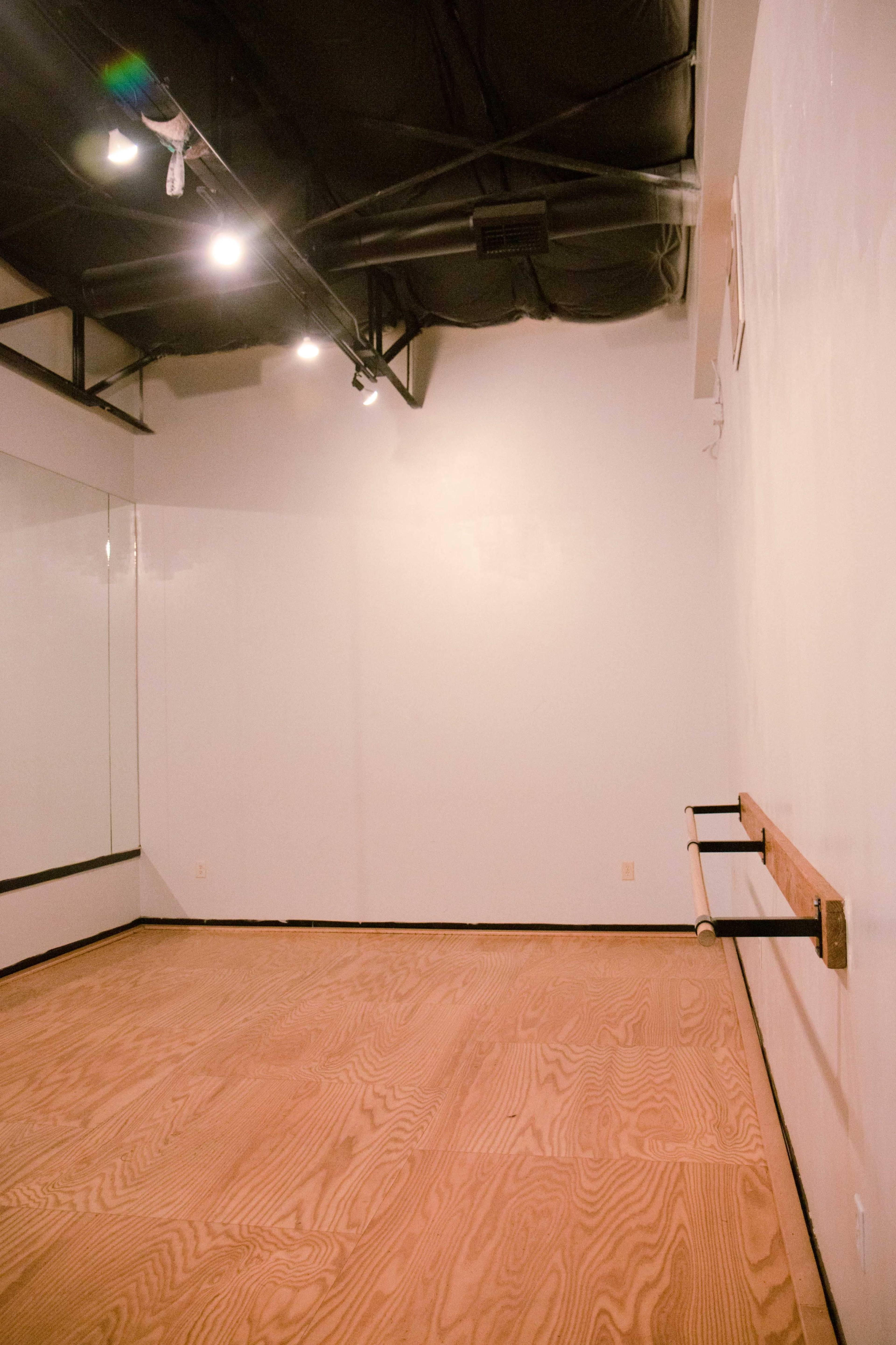 The image shows a dance studio with wooden flooring, a wall-mounted ballet barre, and a mirror partially visible on one wall.