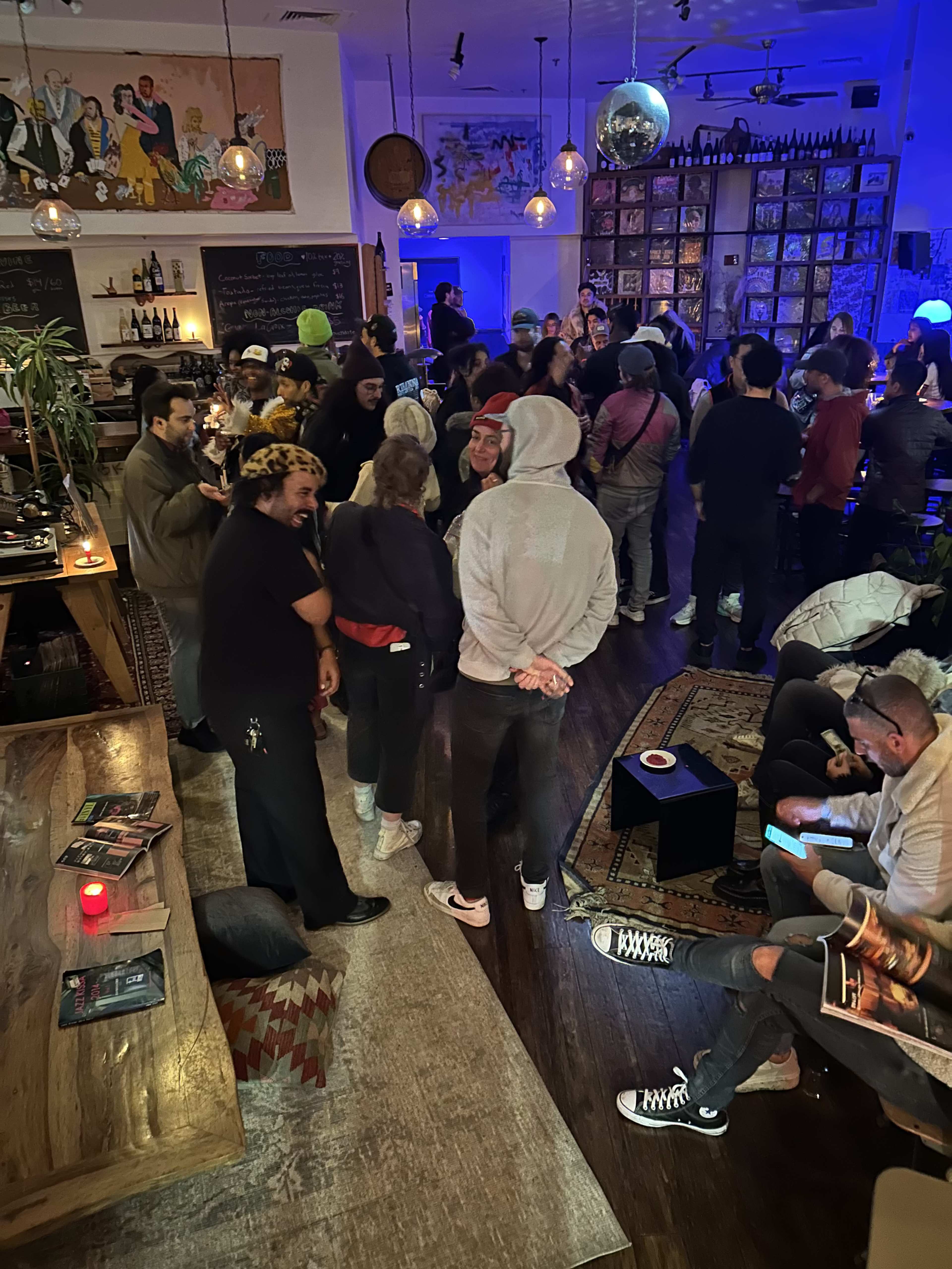 A crowd of people gathers in a dimly lit bar with artwork on the walls and shelves filled with records.