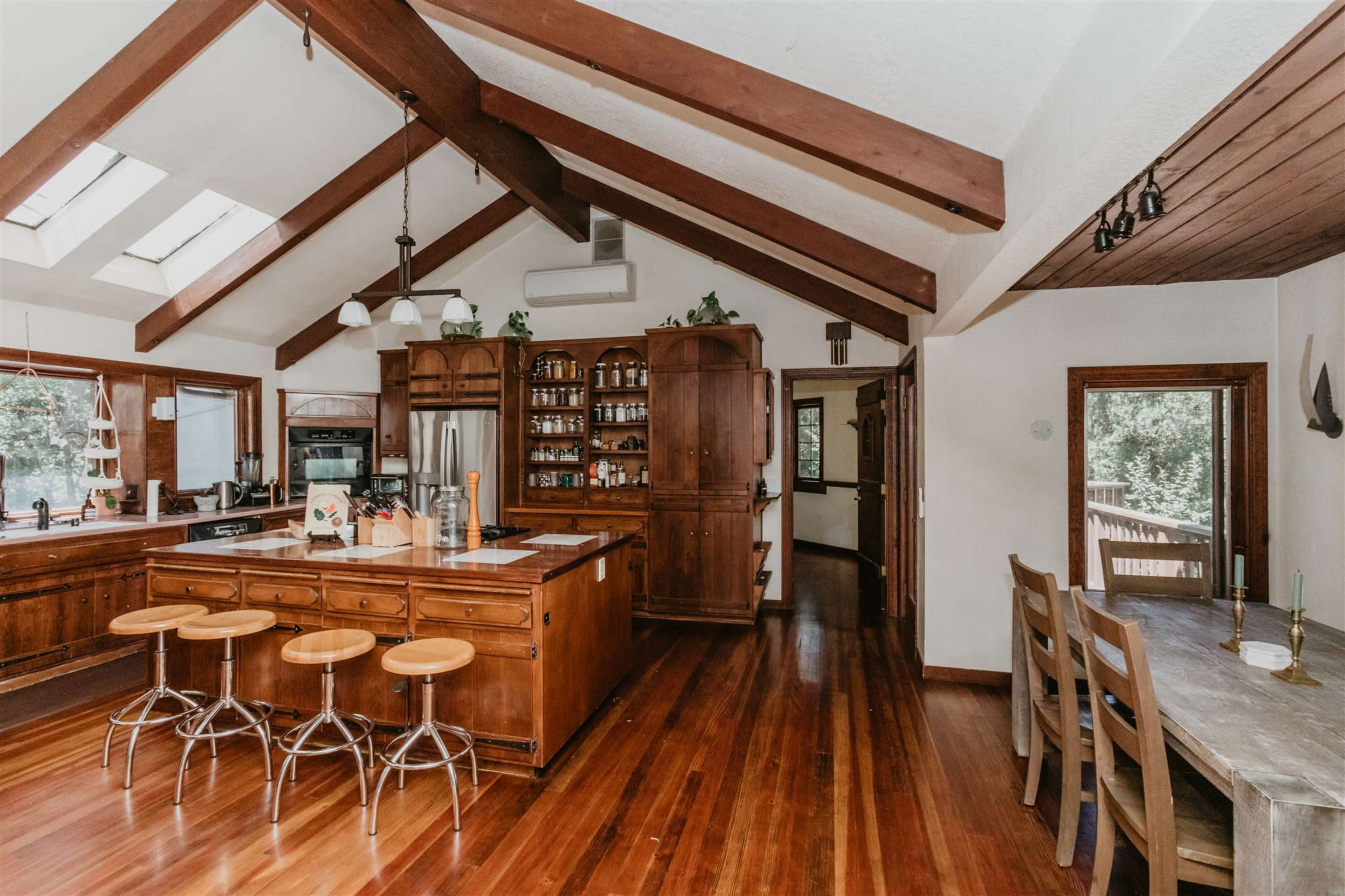 The image shows a spacious kitchen featuring a central island with four wooden stools, vaulted ceiling beams, and natural wood cabinetry.