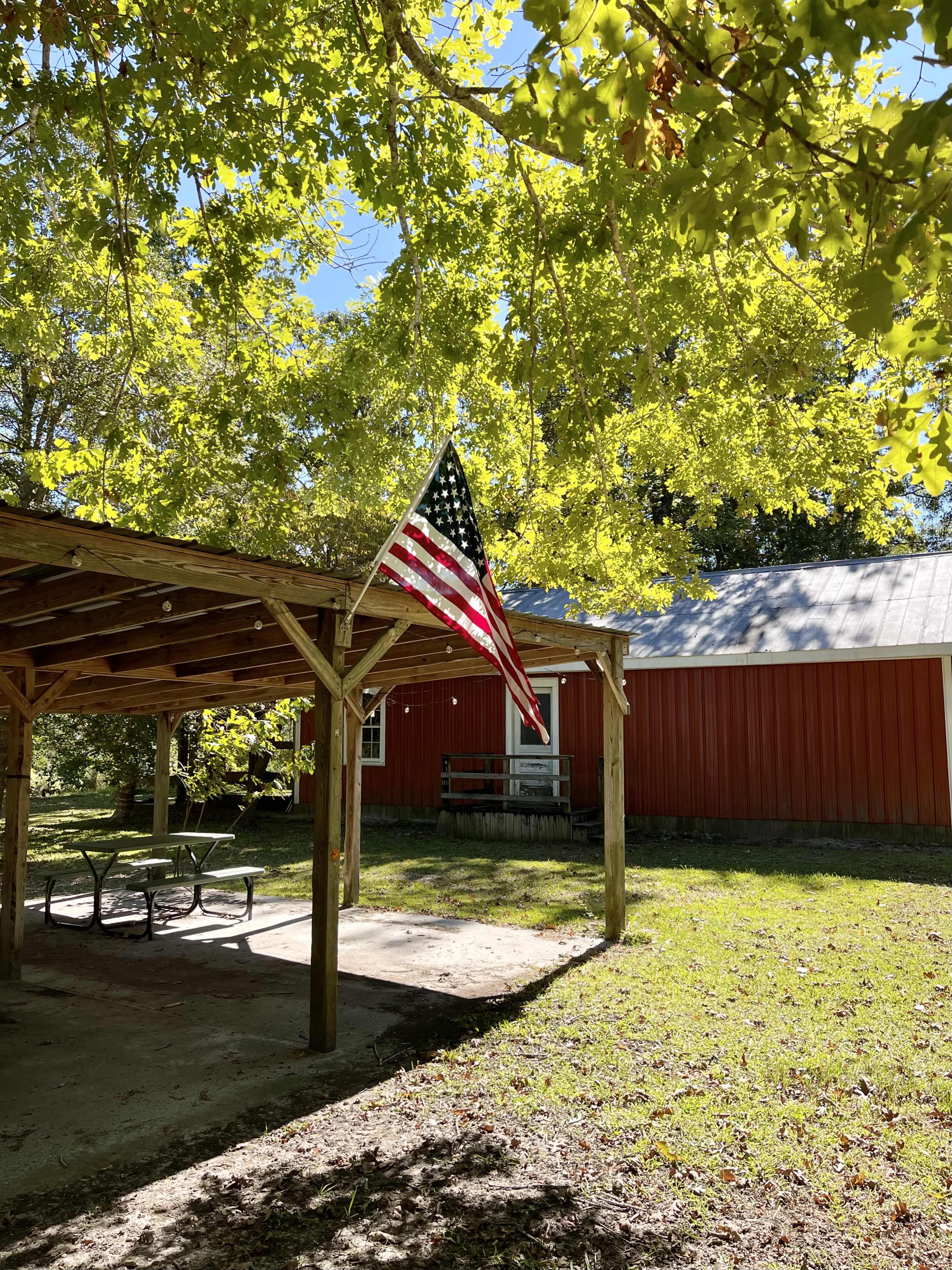 A red building with a white roof is partially visible behind a covered structure, under which an American flag hangs amid green trees.