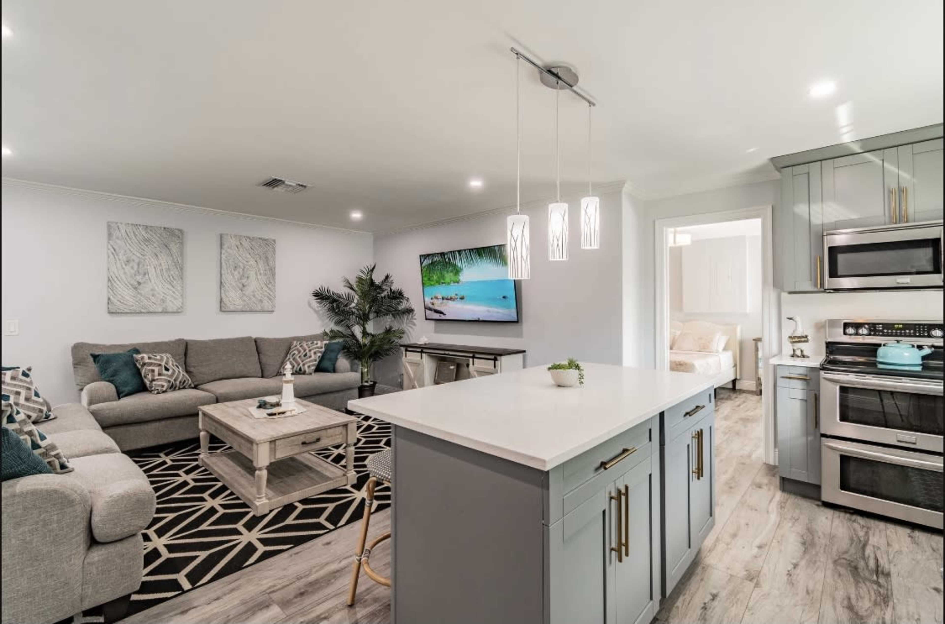 The image shows a modern living space featuring a gray sectional sofa, a coffee table, a wall-mounted TV, and a well-equipped kitchen island.