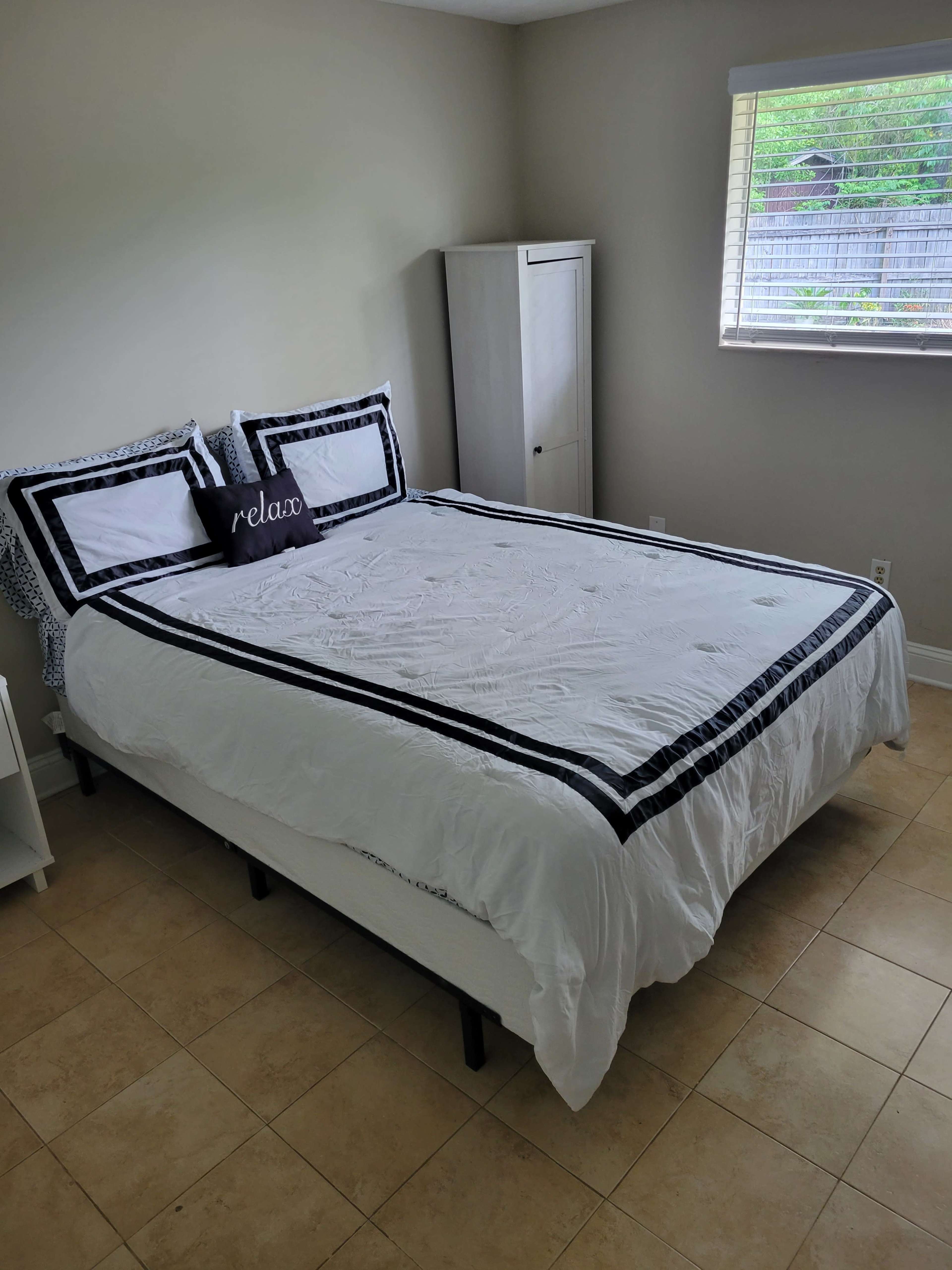 A neatly arranged bedroom features a double bed with a black and white patterned quilt, flanked by two pillows, and a small cabinet against the wall.