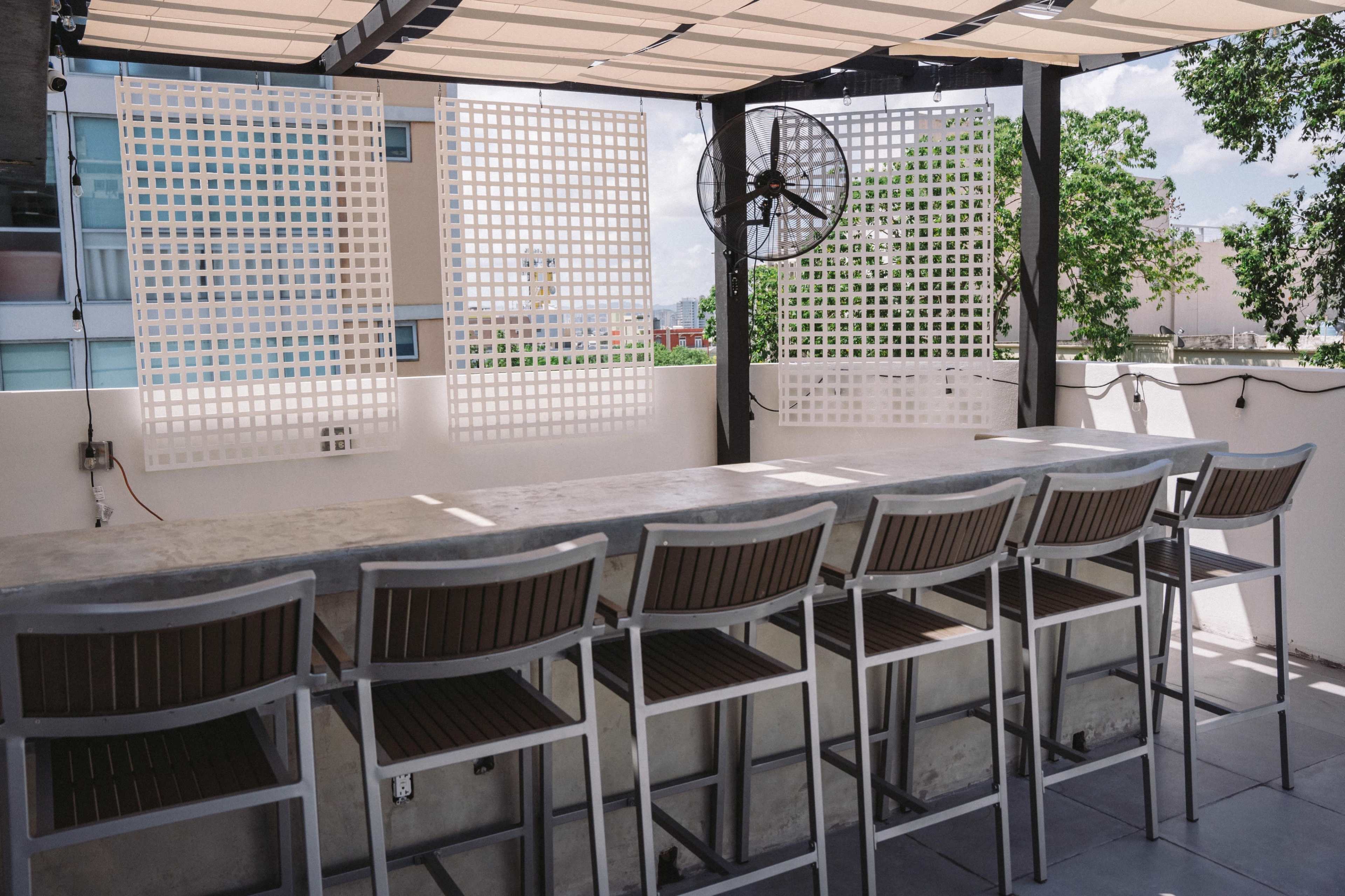 The image shows a rooftop bar area with a long concrete counter and several high stools arranged in front of it, set against a backdrop of white lattice panels and green trees.