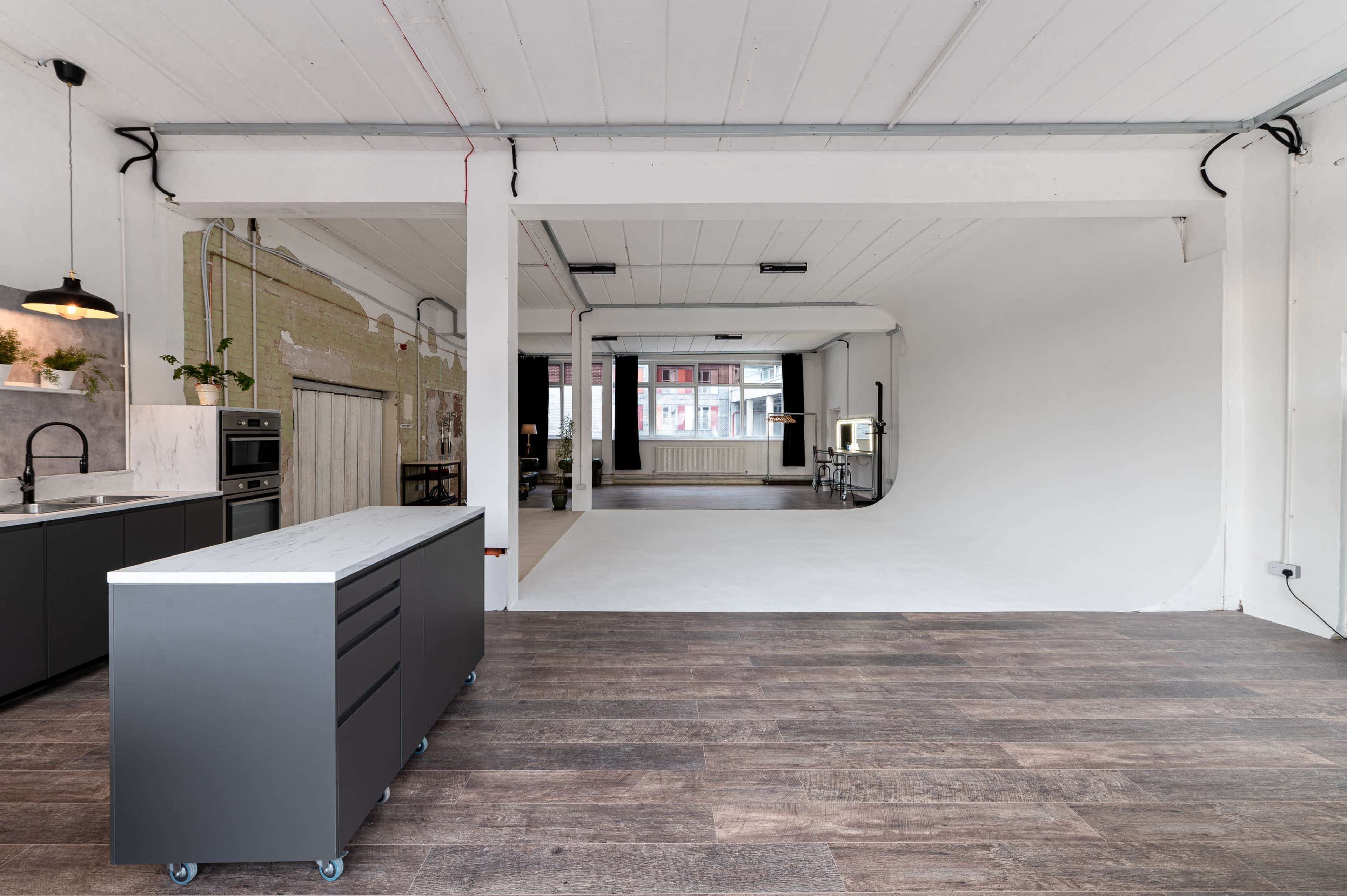 The image shows a spacious, modern studio with a kitchen area on the left and an empty white backdrop on the right.