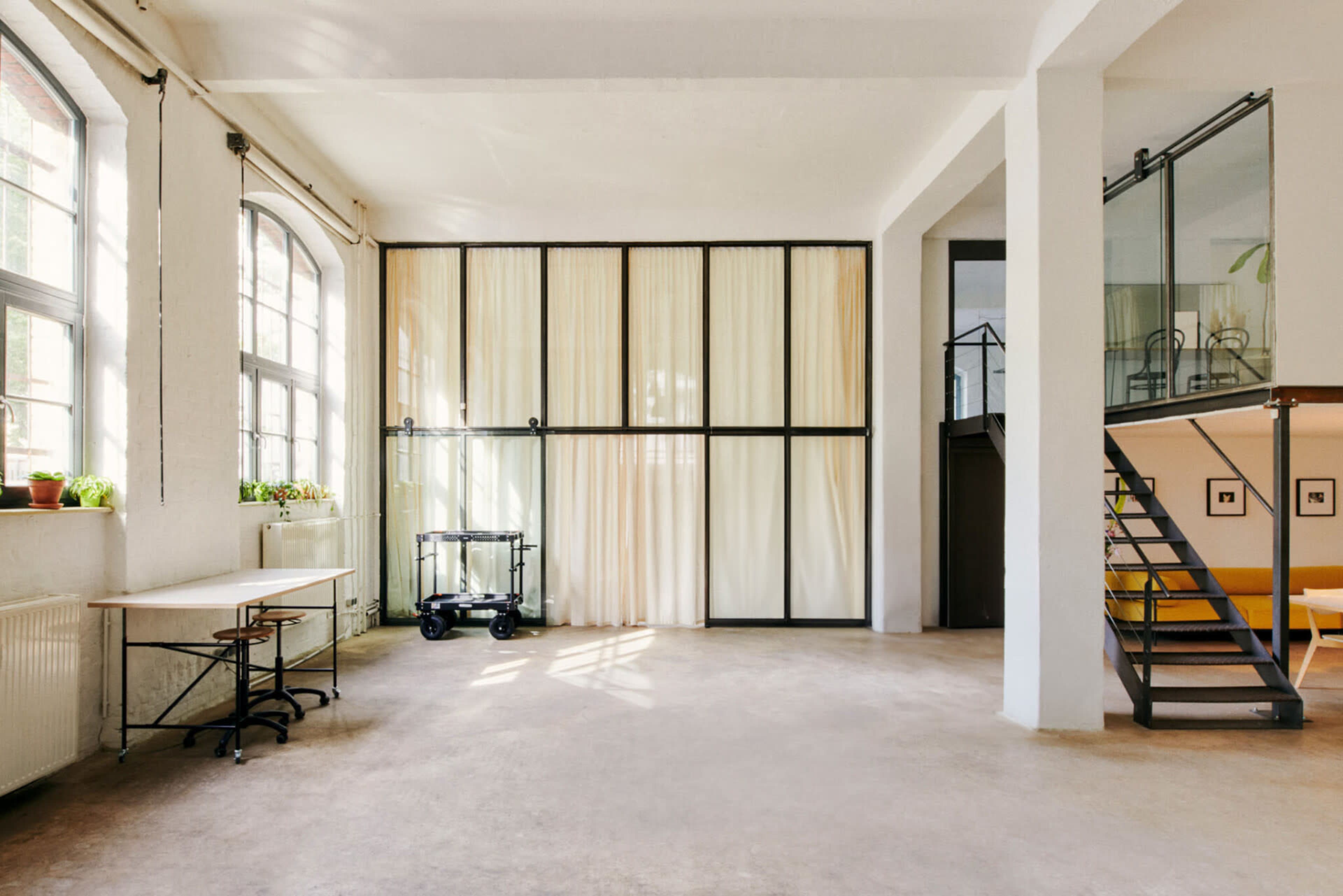 A spacious, well-lit room features large windows, a curtain wall, and a staircase leading to a loft area.
