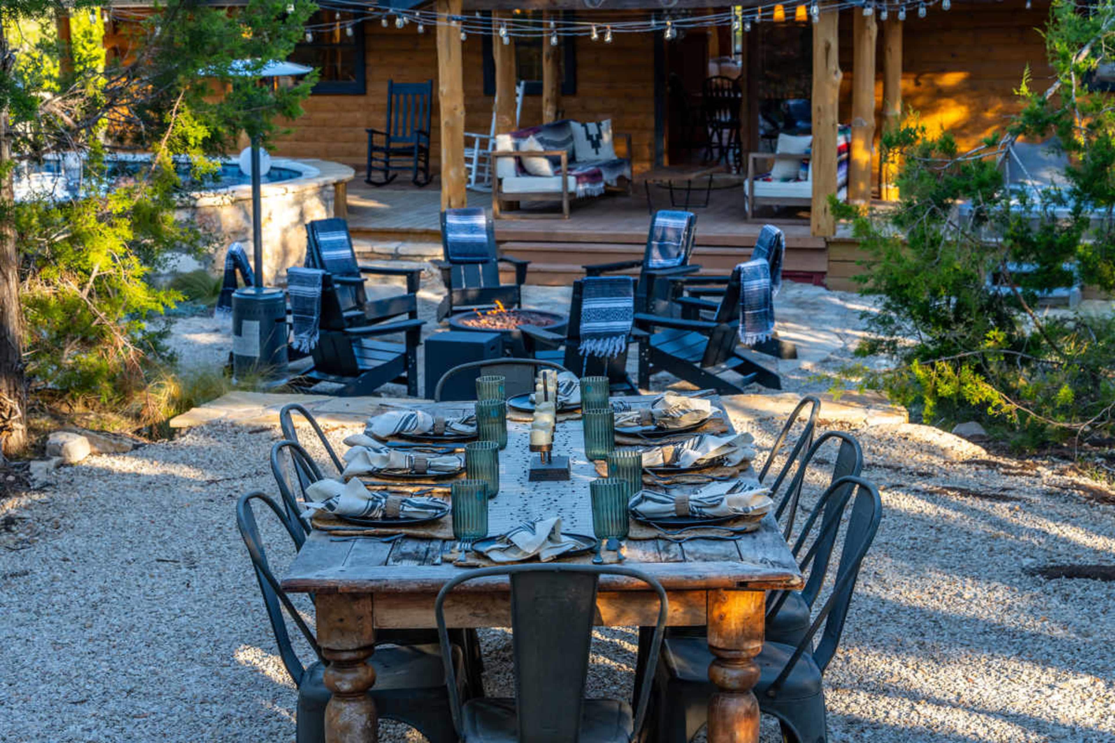 A rustic wooden dining table is set with plates and glasses, surrounded by metal chairs in an outdoor space with a cozy cabin in the background.