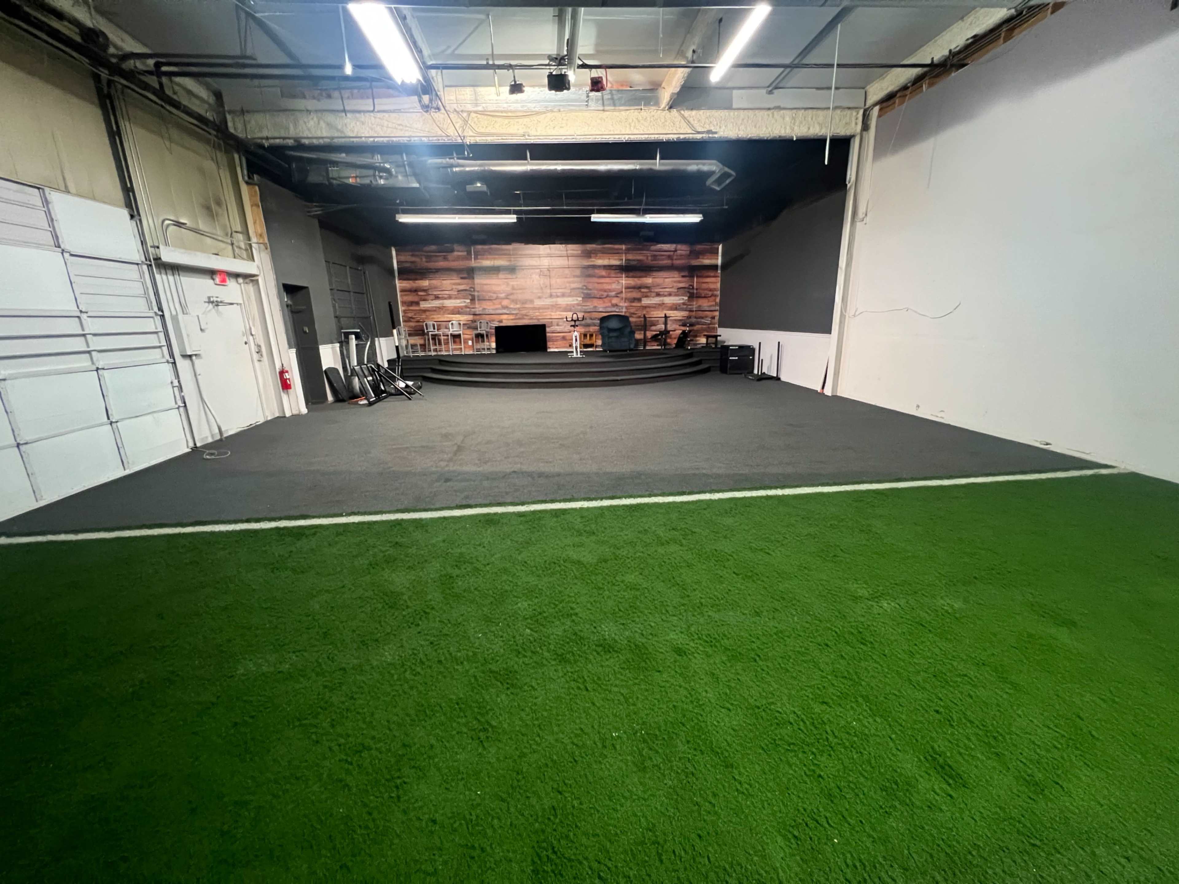 The image shows a spacious indoor area with a green turf section and a raised platform at the back featuring wooden paneling and a chair.