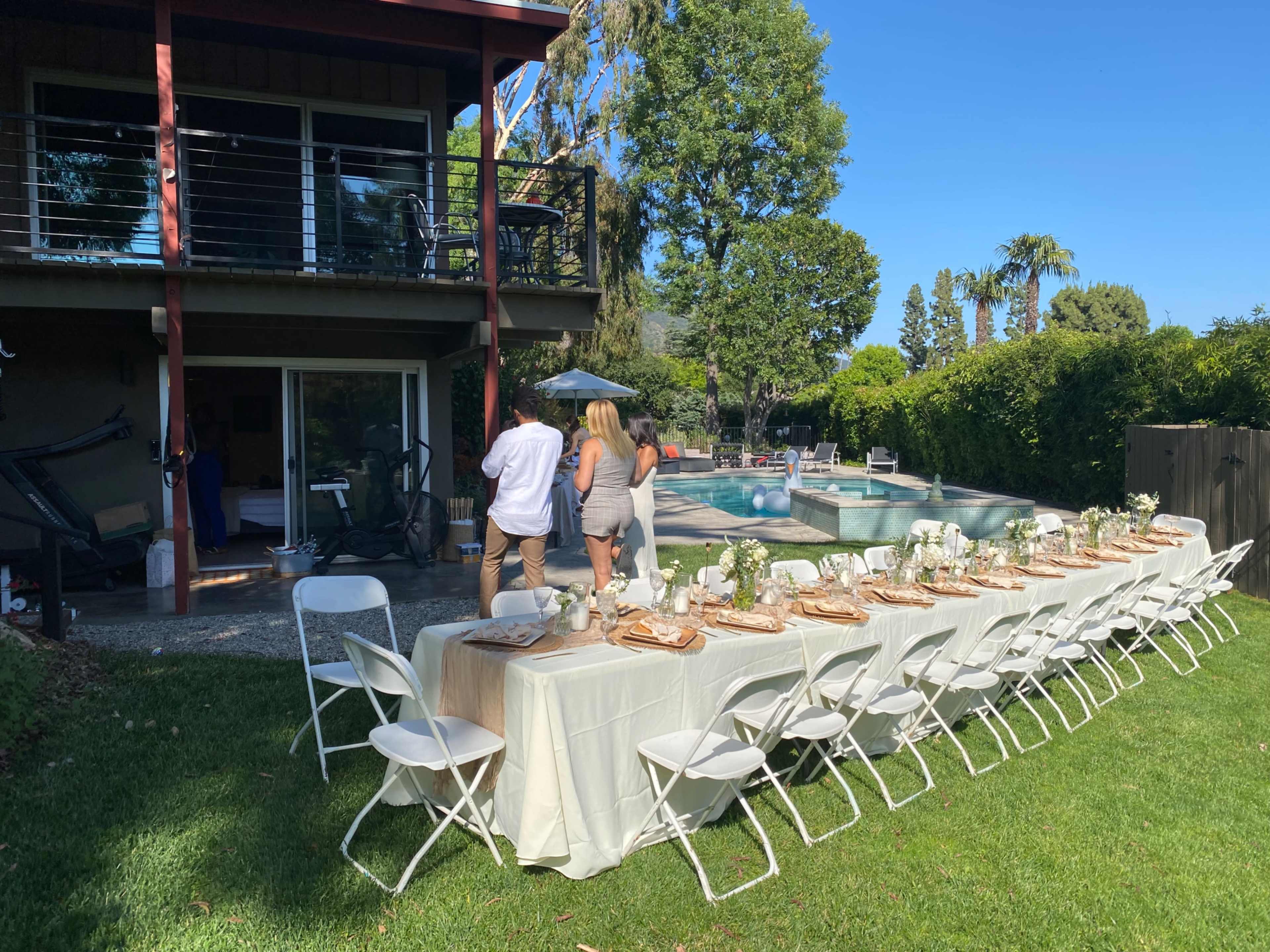 A long dining table is set up outdoors near a pool, with guests walking towards it while surrounded by greenery and a two-story house in the background.