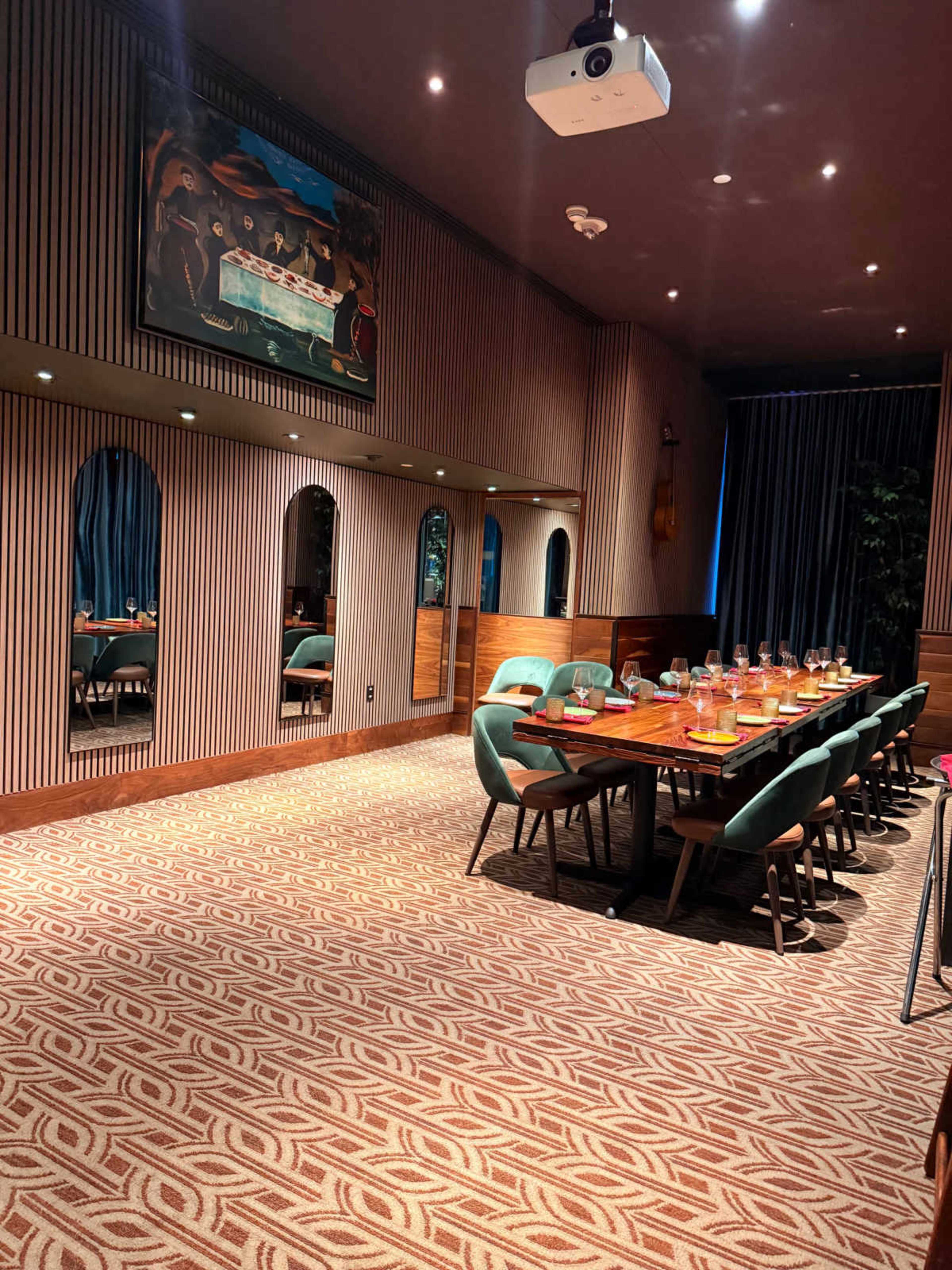 The image depicts a modern dining room with a long table set for a meal, surrounded by chairs, and featuring patterned carpeting and decorative mirrors on the walls.