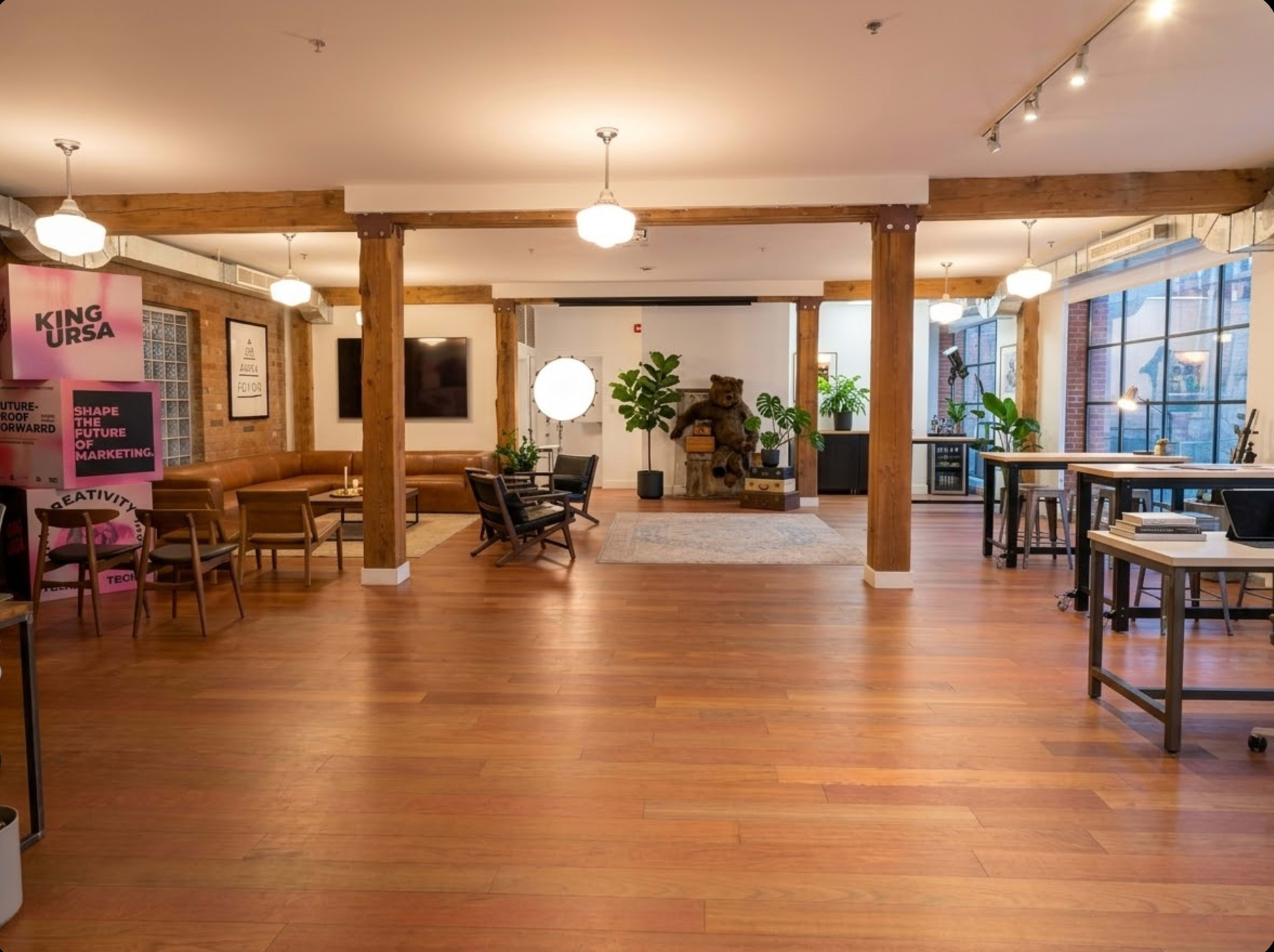 The image shows a spacious, well-lit office space with wooden floors, decorative potted plants, and seating areas arranged around a central gathering space.