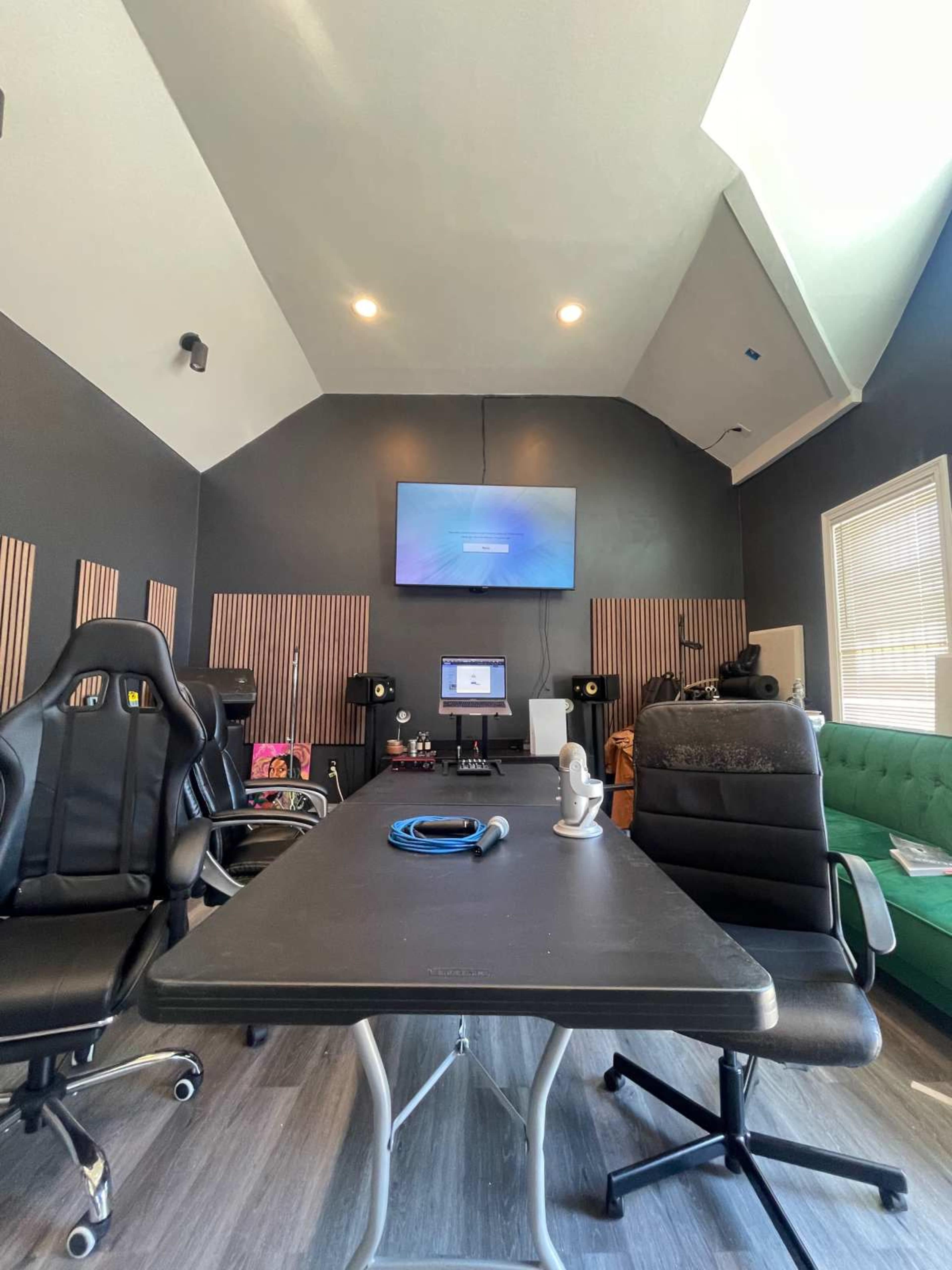The image shows a recording studio with a large table, two black chairs, a computer setup, and acoustic panels on the walls.