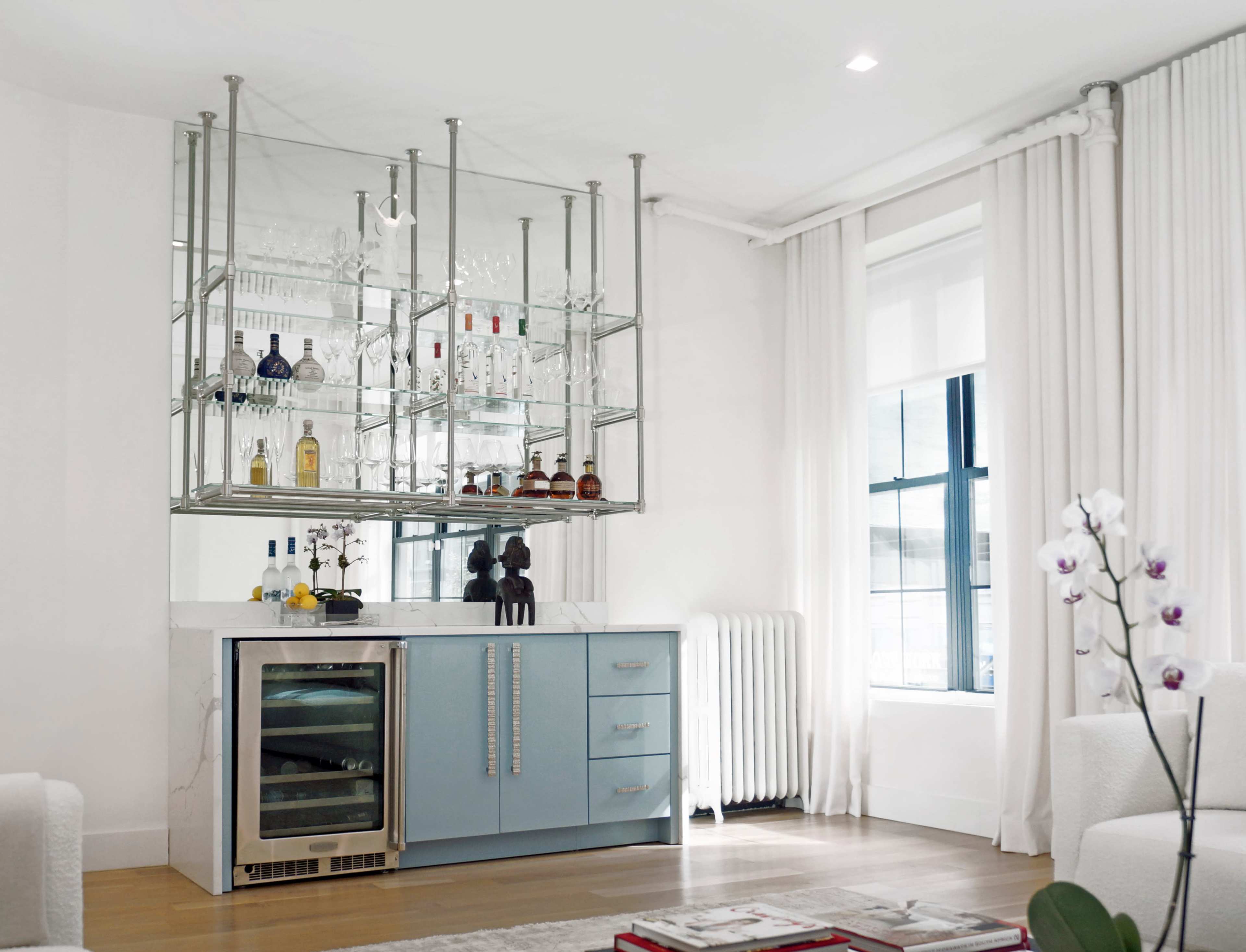 The image shows a modern home bar setup with a sleek, light blue cabinet and a glass rack displaying various bottles and glasses above it.