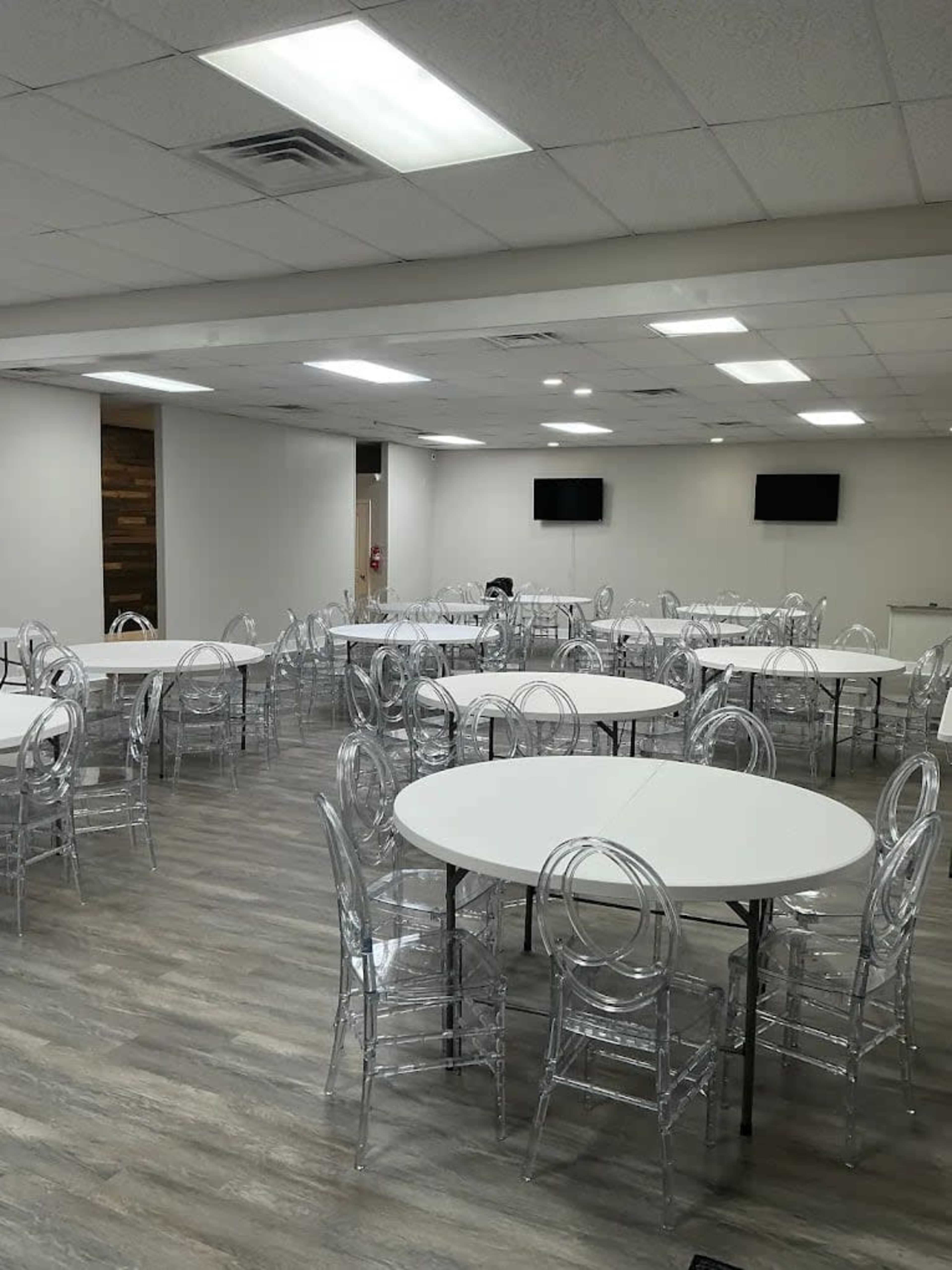 The image shows a large, empty room with multiple circular tables and transparent chairs, illuminated by overhead lighting.