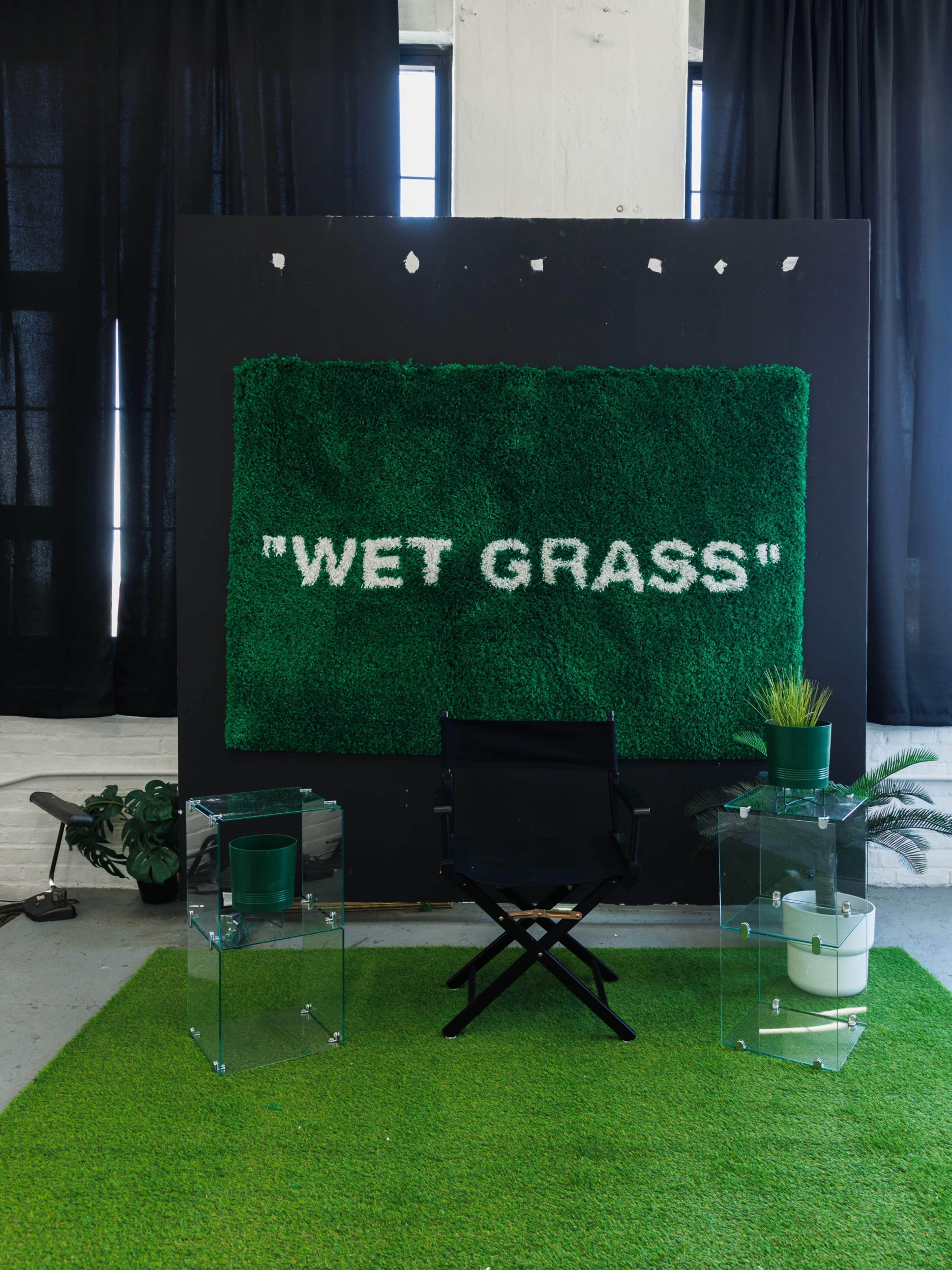 The scene features a green artificial grass wall with the words "WET GRASS" displayed in white, flanked by a black director's chair and two clear acrylic side tables with potted plants on them.