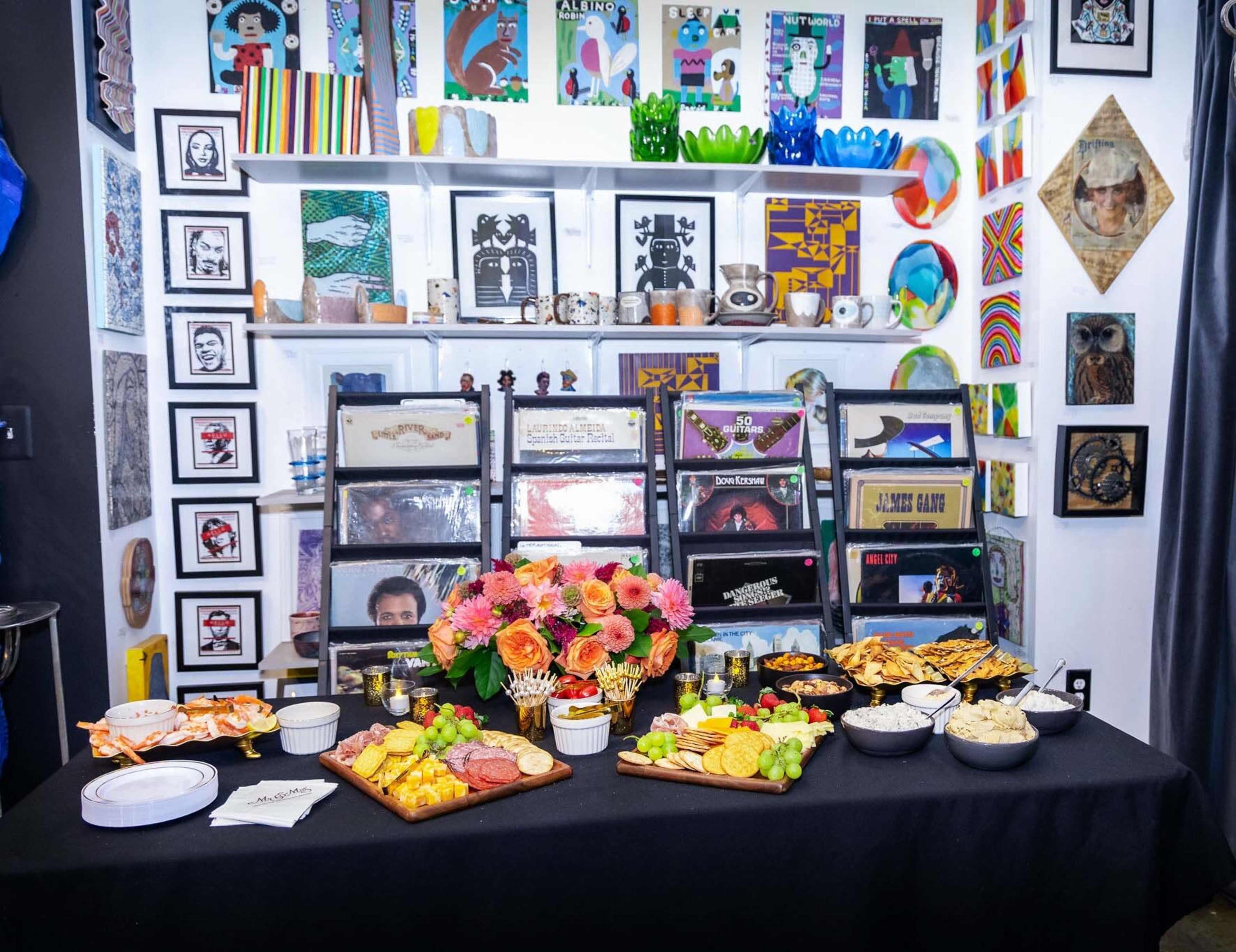A table covered with various platters of food is set against a backdrop of colorful artwork and framed pictures on a wall.