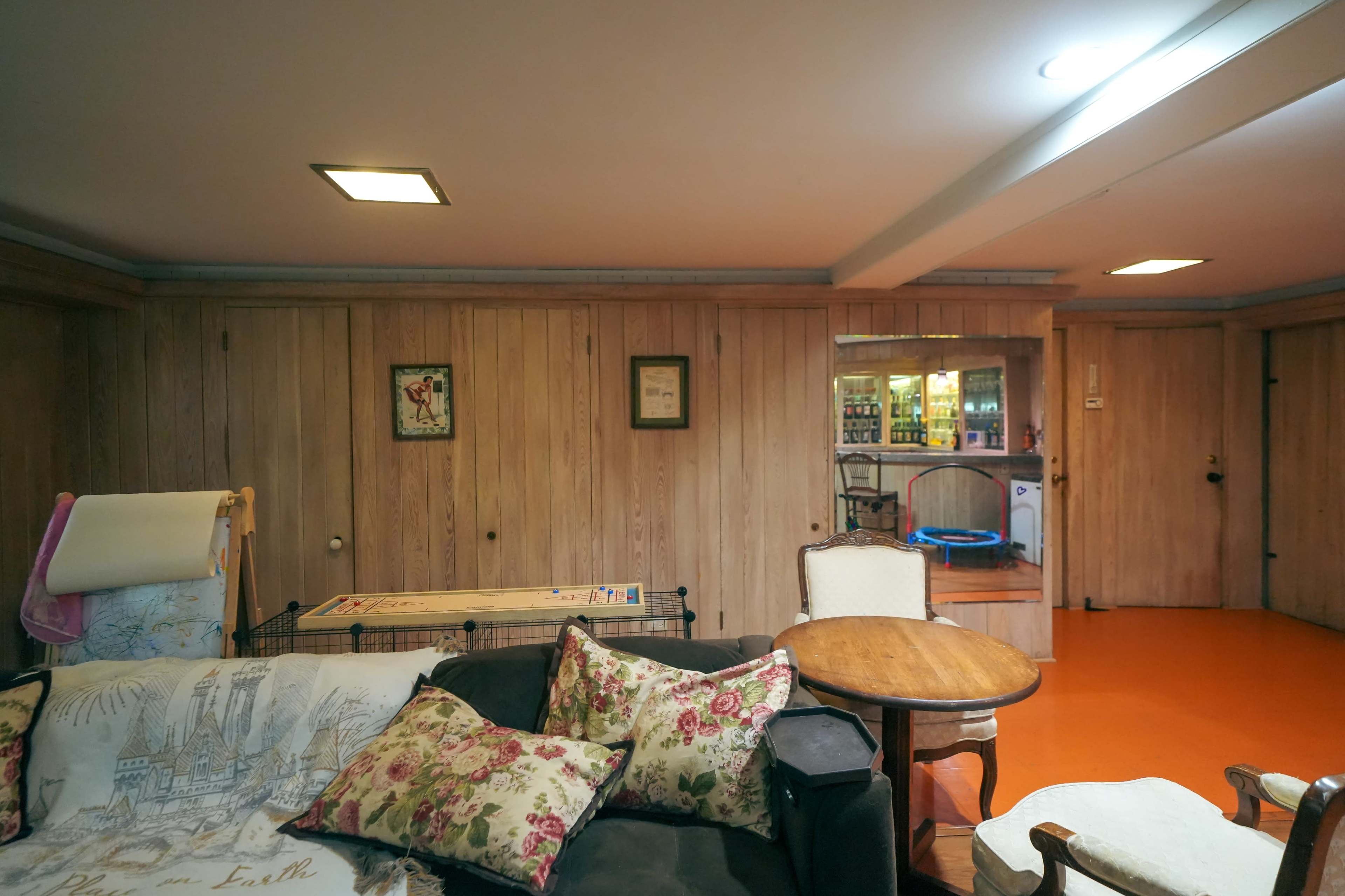 A basement room with wooden paneling, a sofa with floral pillows, a round table, and a visible kitchenette area in the background.
