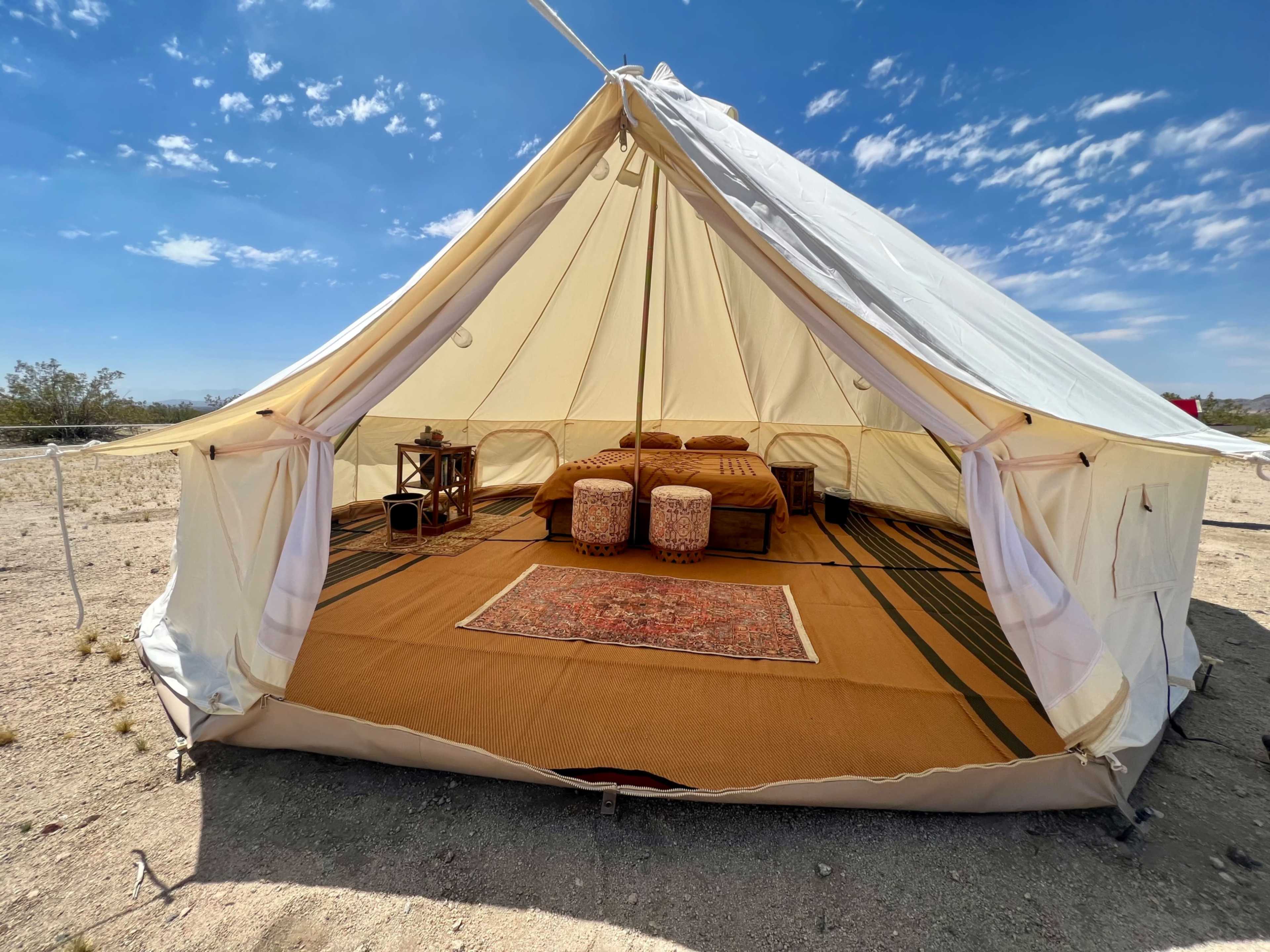 A spacious, cream-colored canvas tent is set up in a desert landscape, featuring a neatly arranged interior with a bed, two poufs, and a decorative rug.