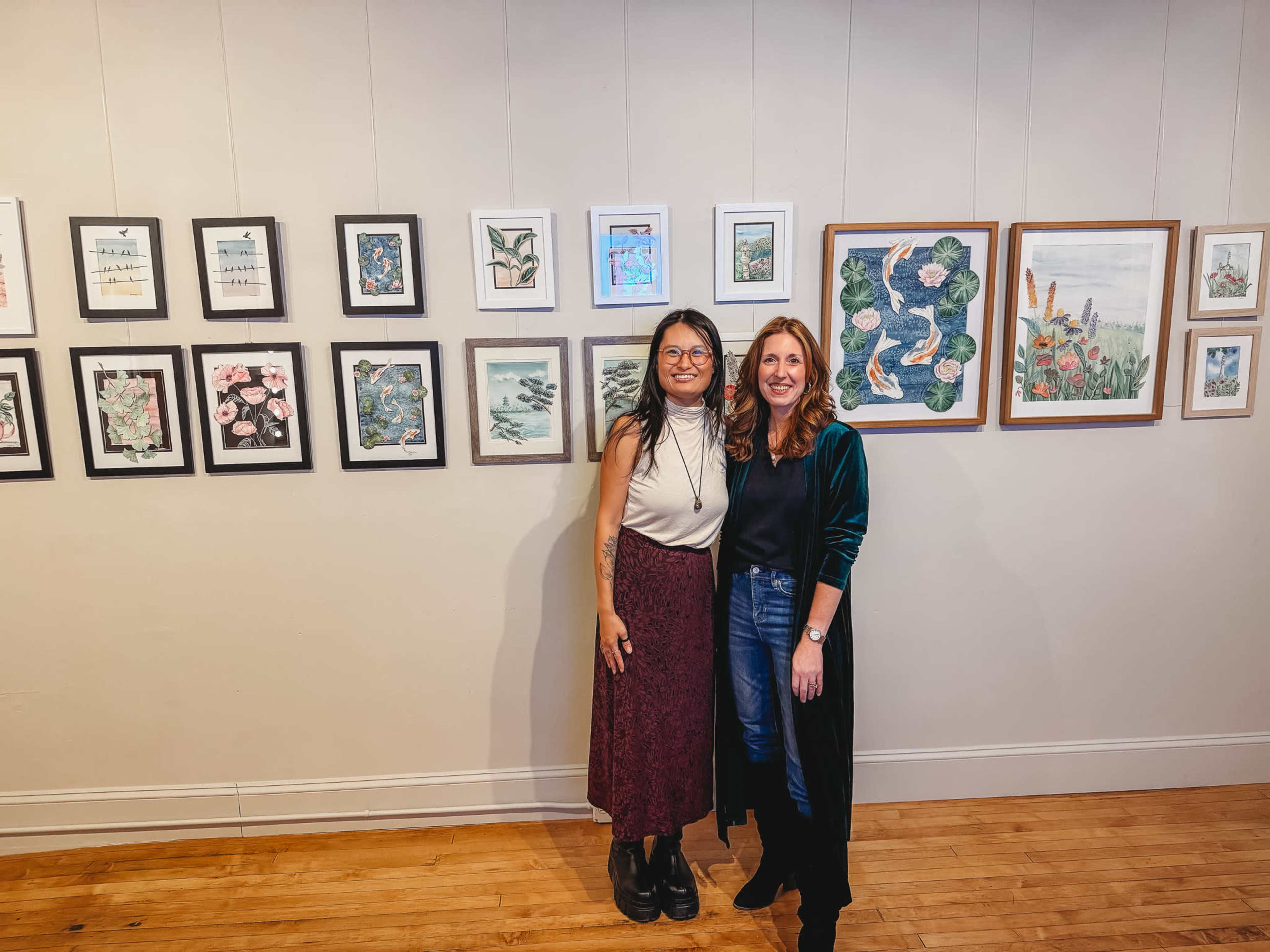 Two women stand together in an art gallery, smiling in front of a collection of framed artworks hanging on the wall behind them.