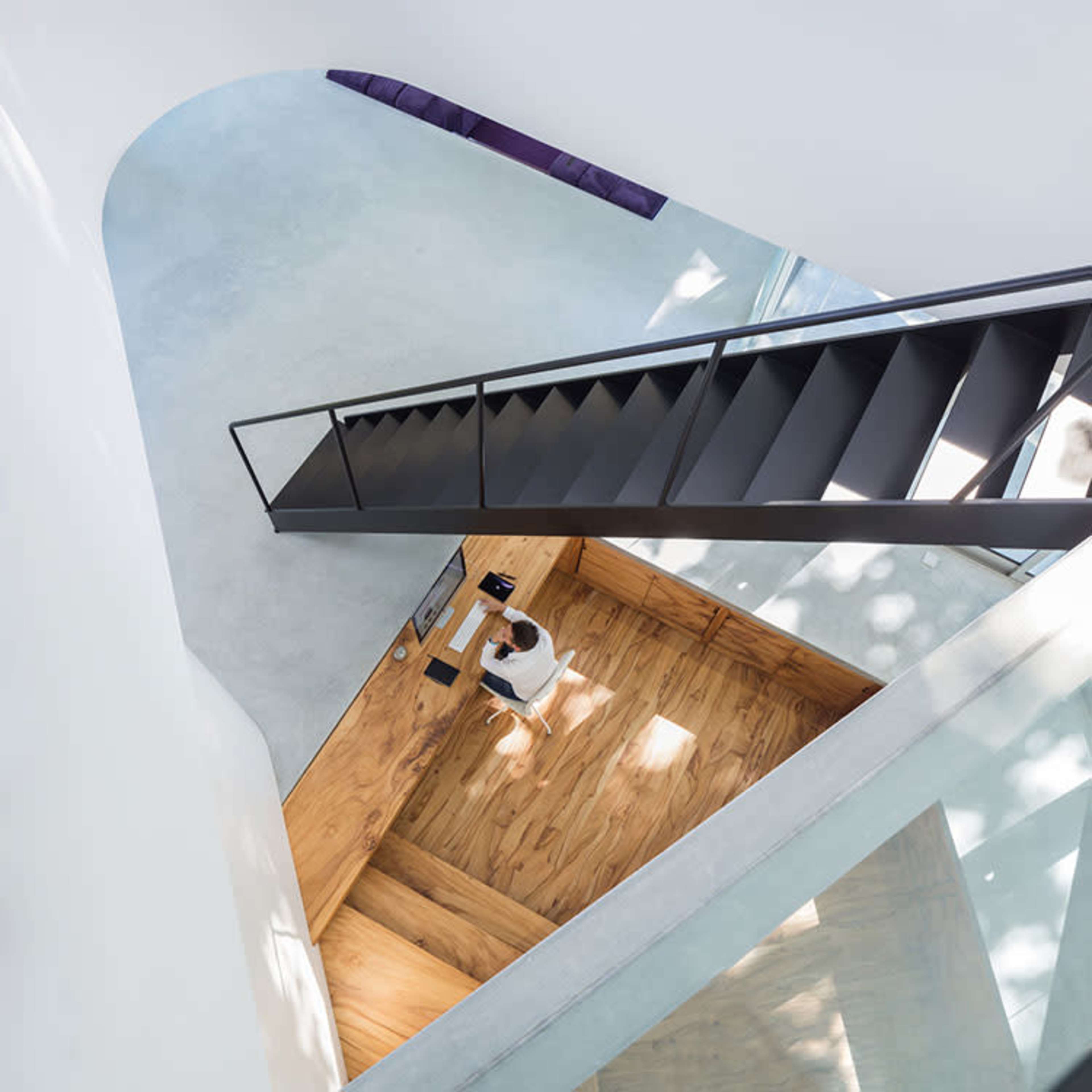 A person stands at a modern wooden desk in a spacious interior with an open staircase and minimalist design.