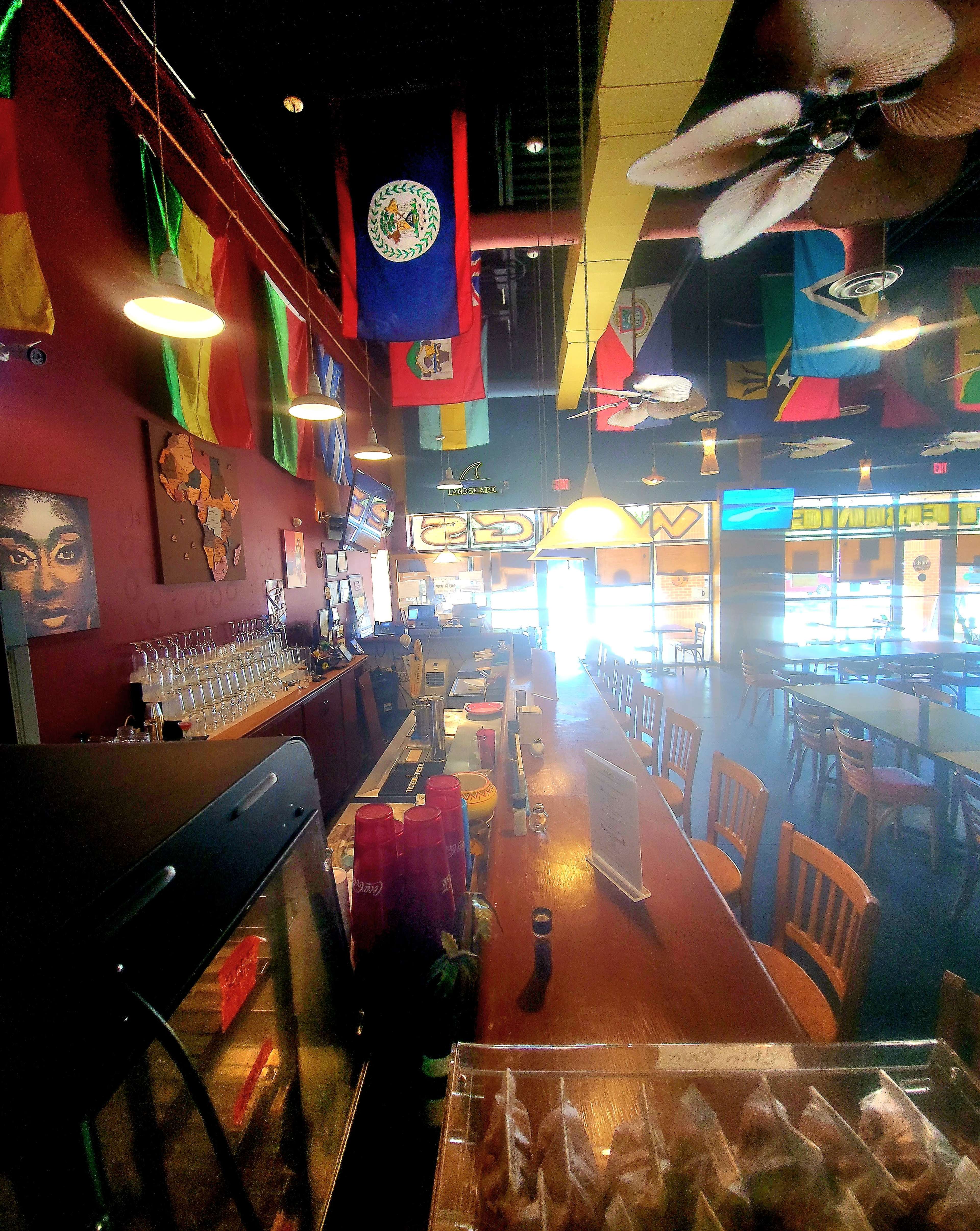 The image shows a bar area in a restaurant decorated with various international flags hanging from the ceiling, and an organized counter with bar stools and glassware on display.
