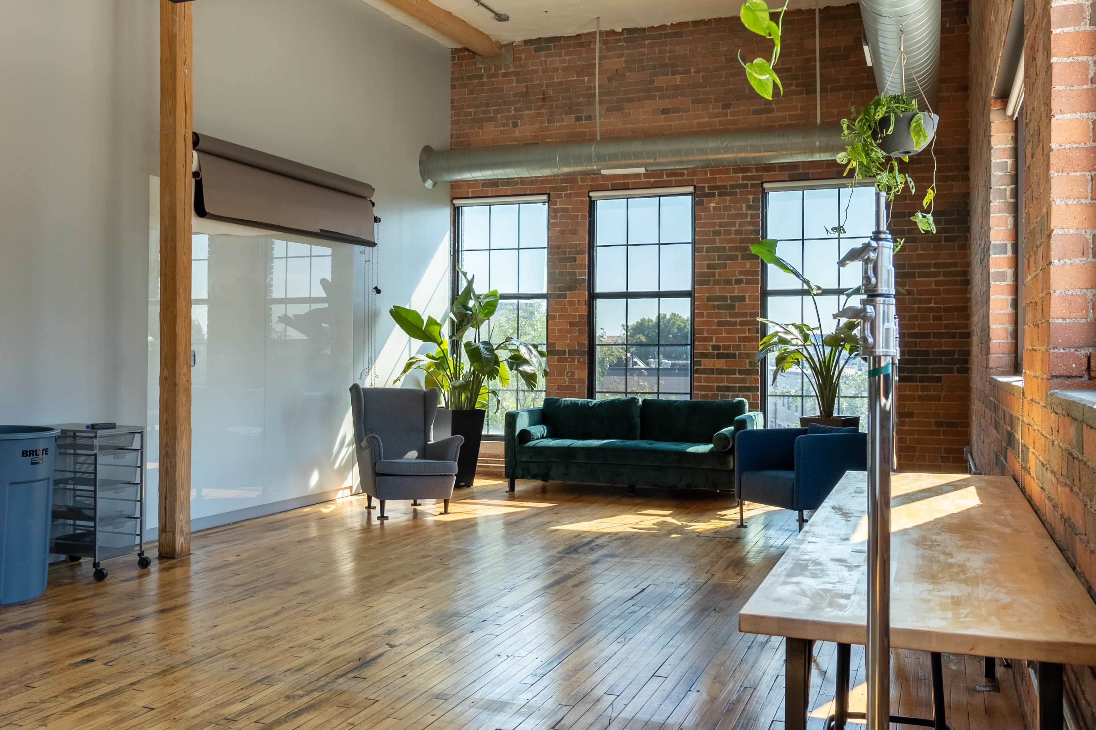 A spacious, well-lit room with brick walls, large windows, a green sofa, an armchair, and potted plants.