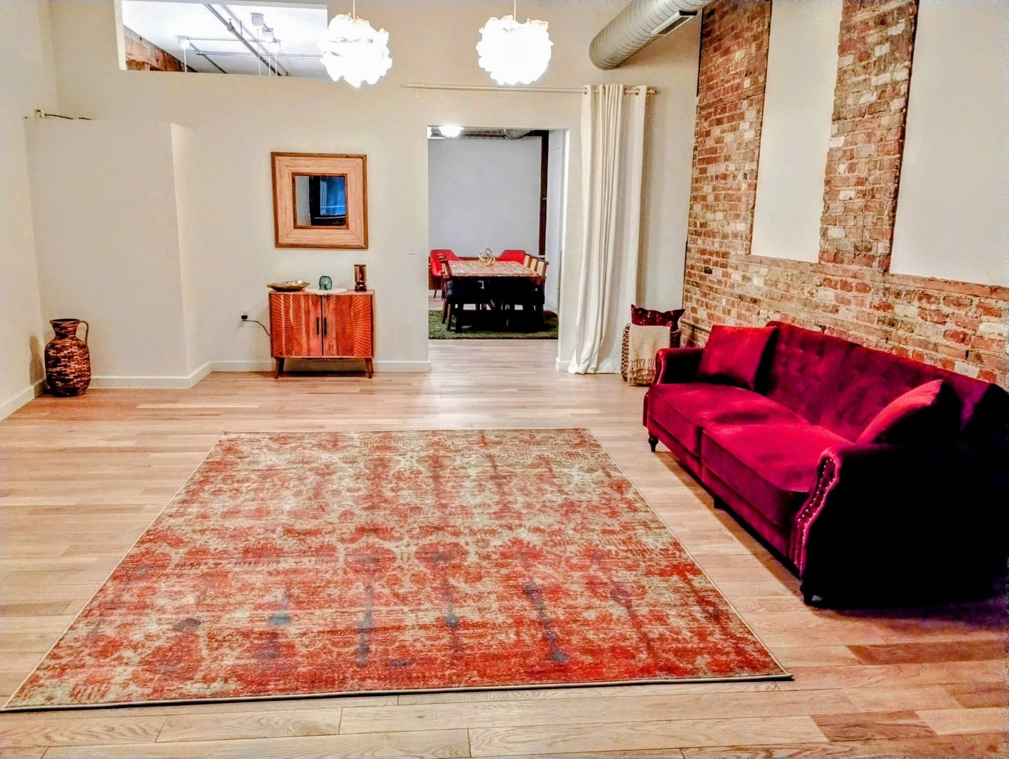 The image shows a spacious interior with a wooden floor, a large patterned rug, a red sofa, and a decorative wooden cabinet against a brick wall, leading to a dining area in the background.