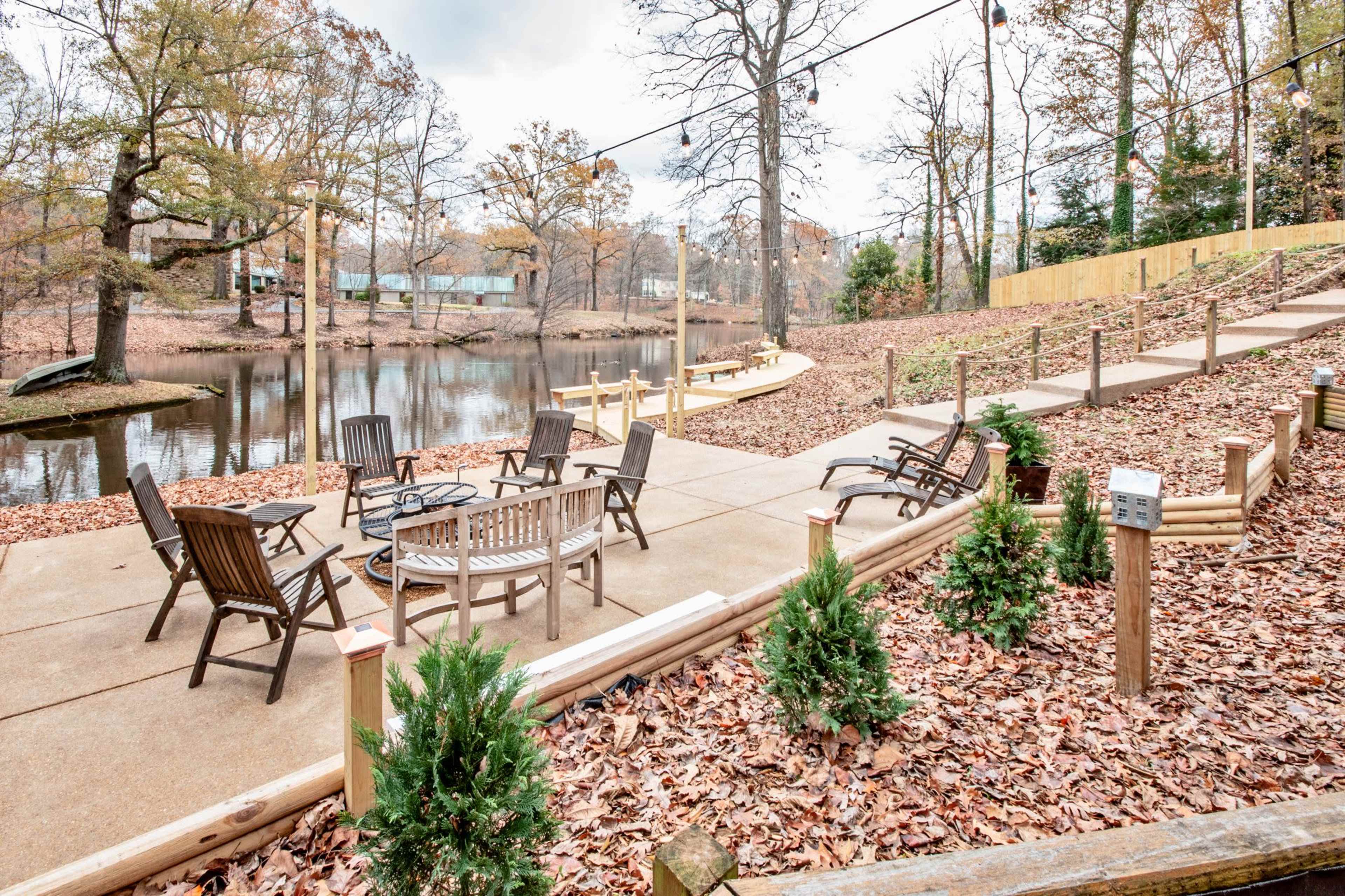A patio area by a pond, featuring multiple wooden chairs and tables arranged on a concrete surface, surrounded by fallen leaves and small shrubbery.