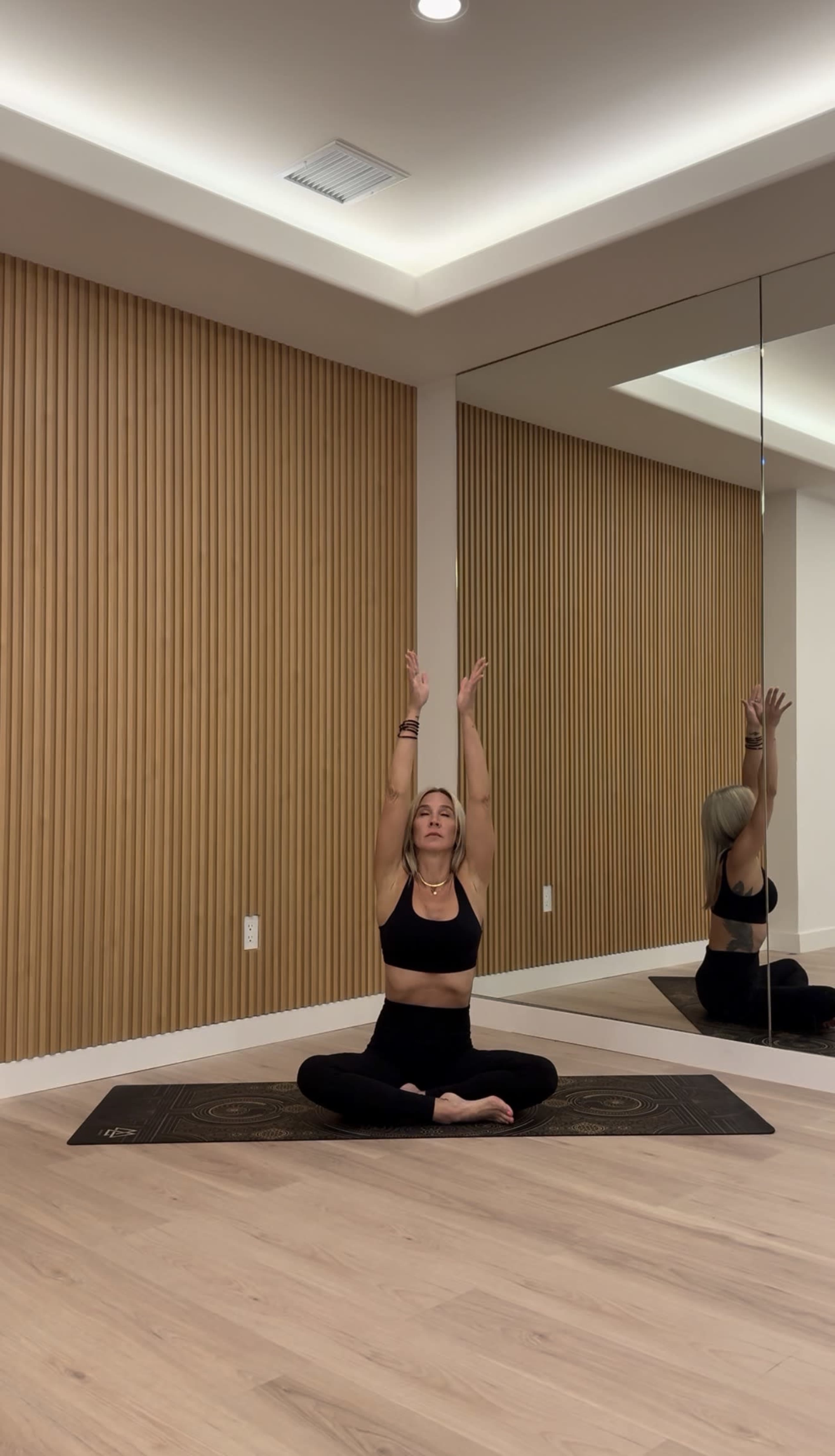 A person is seated in a yoga pose on a mat in a room with wooden paneling and a mirror.