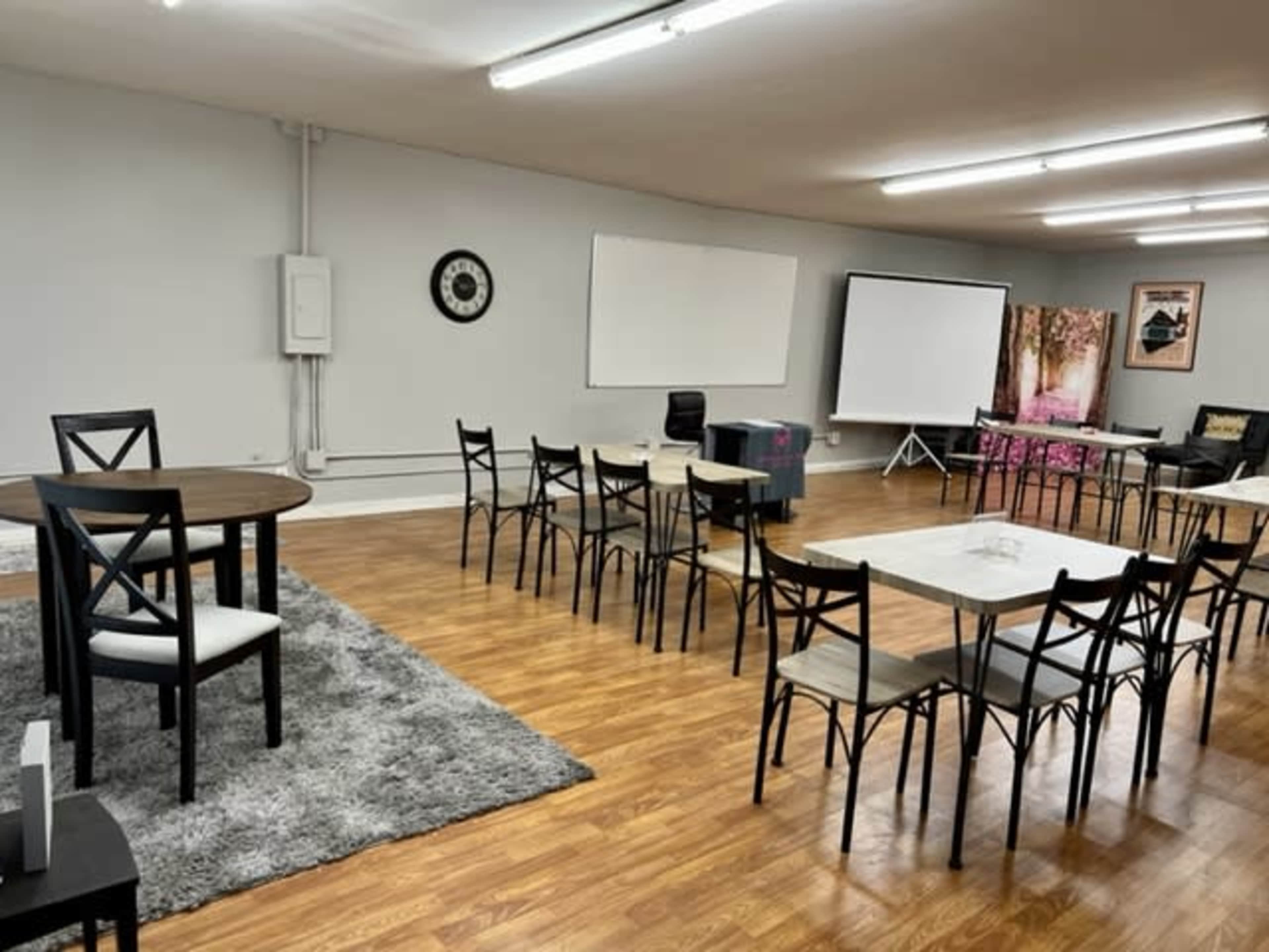 A spacious meeting room features a series of tables and chairs arranged in a circle, with a projector screen and whiteboards on opposite walls.