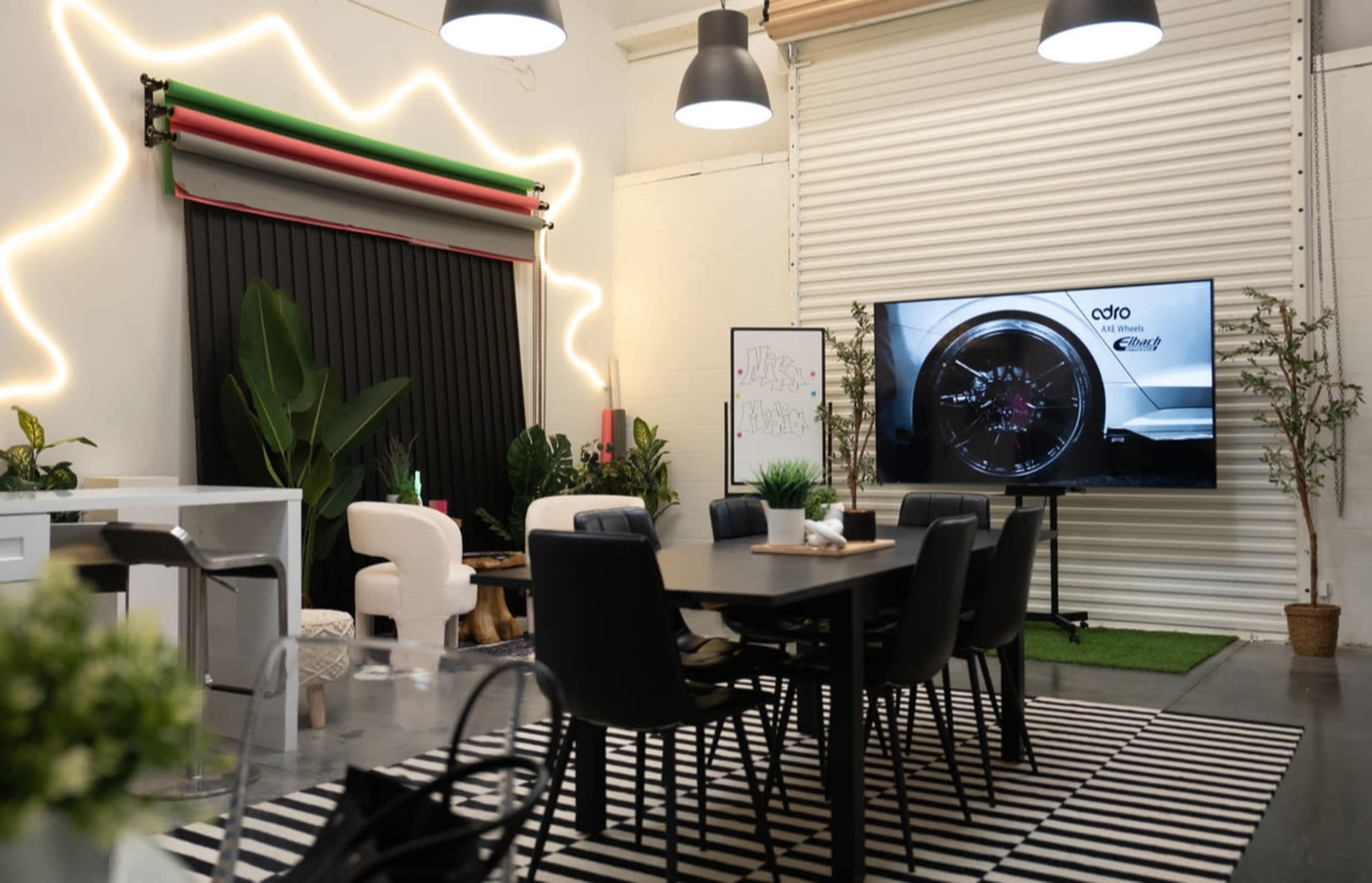 The image shows a modern indoor workspace with a large screen displaying a close-up of a car wheel, surrounded by sleek furniture, plants, and decorative lighting.