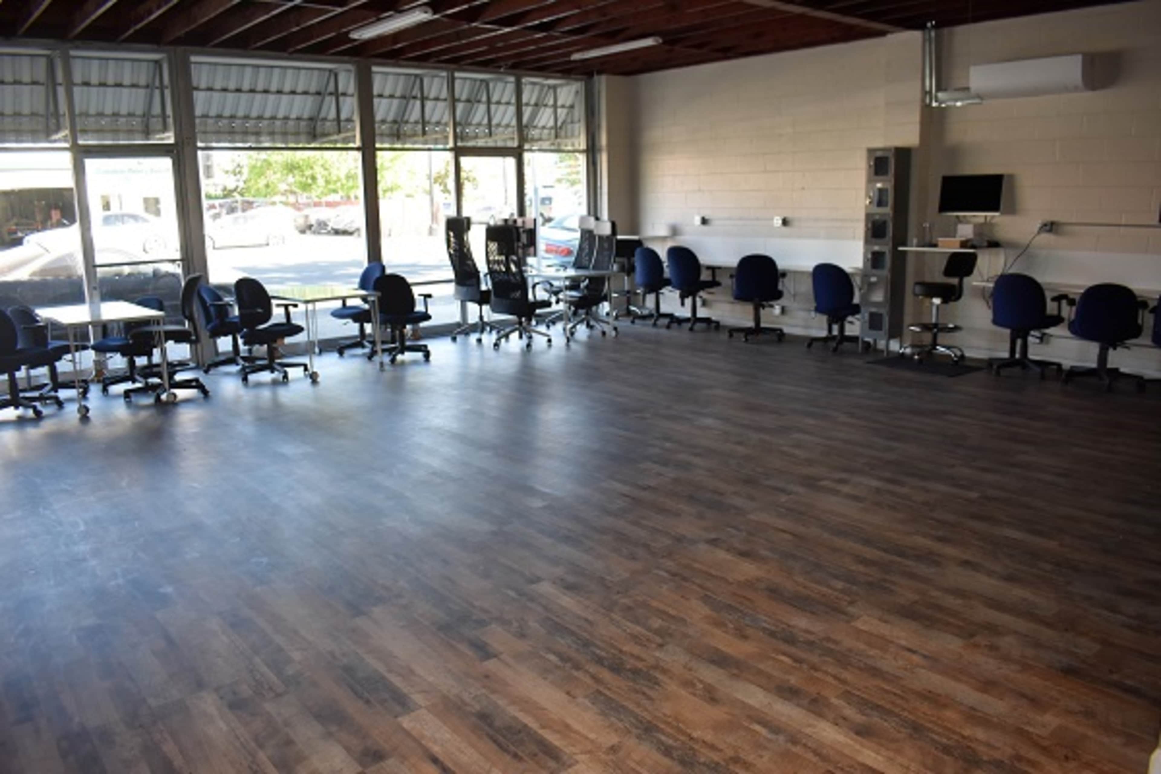 An empty room with wooden flooring, lined with office chairs and tables, and large windows letting in natural light.