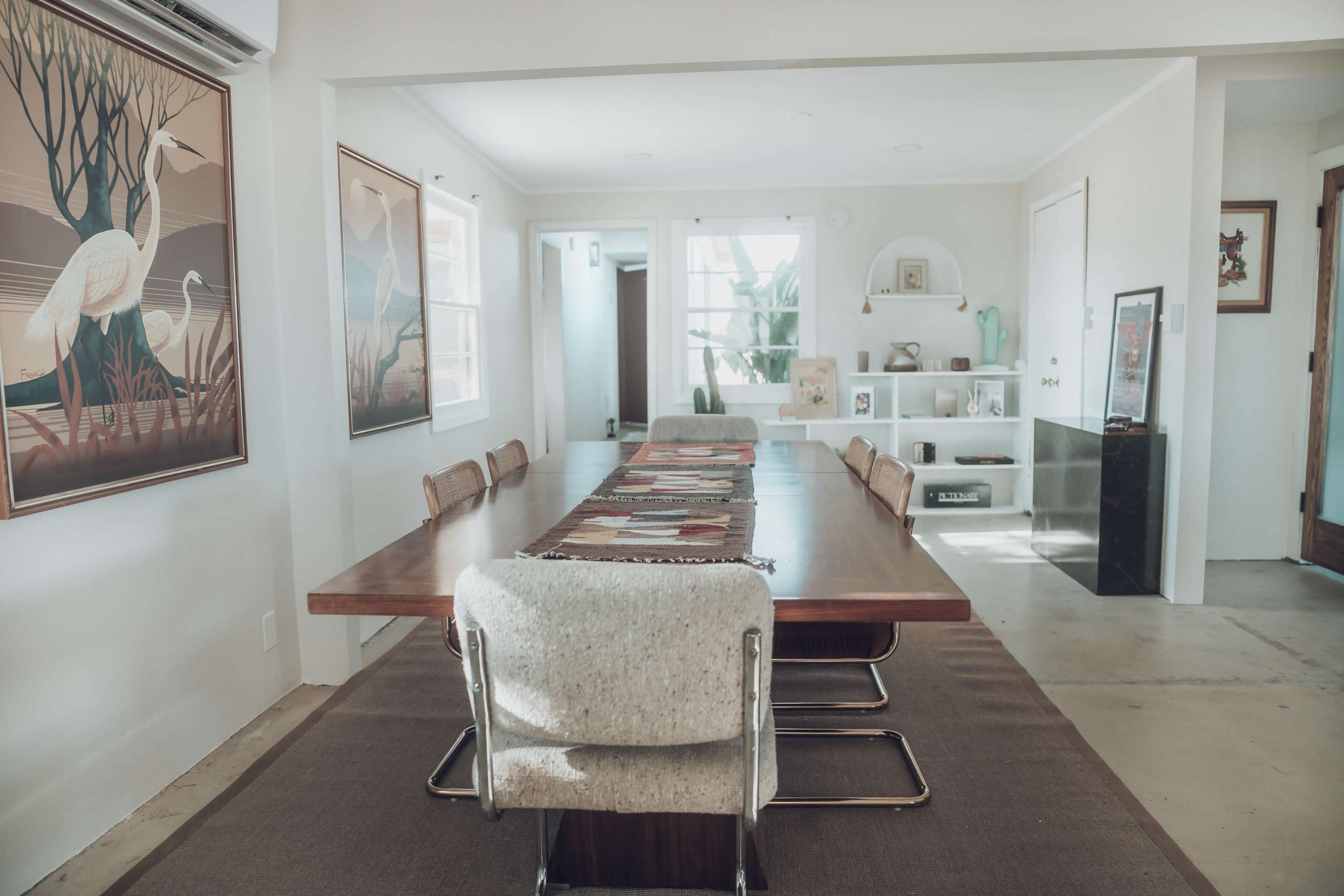 A modern dining room features a long wooden table surrounded by upholstered chairs, with artwork on the walls and a shelving unit in the background.