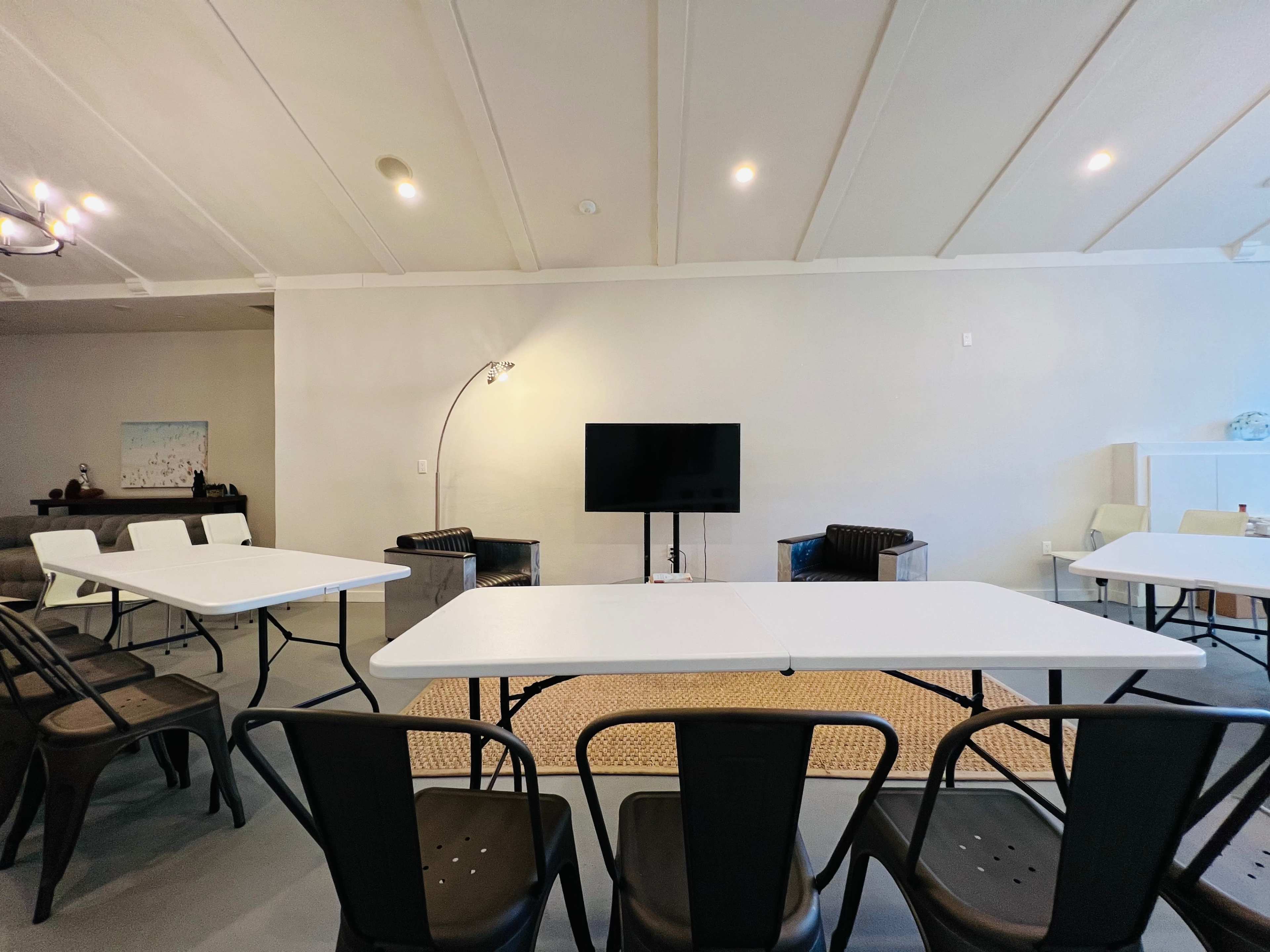 A spacious meeting room with multiple white tables and black chairs arranged around a large television.