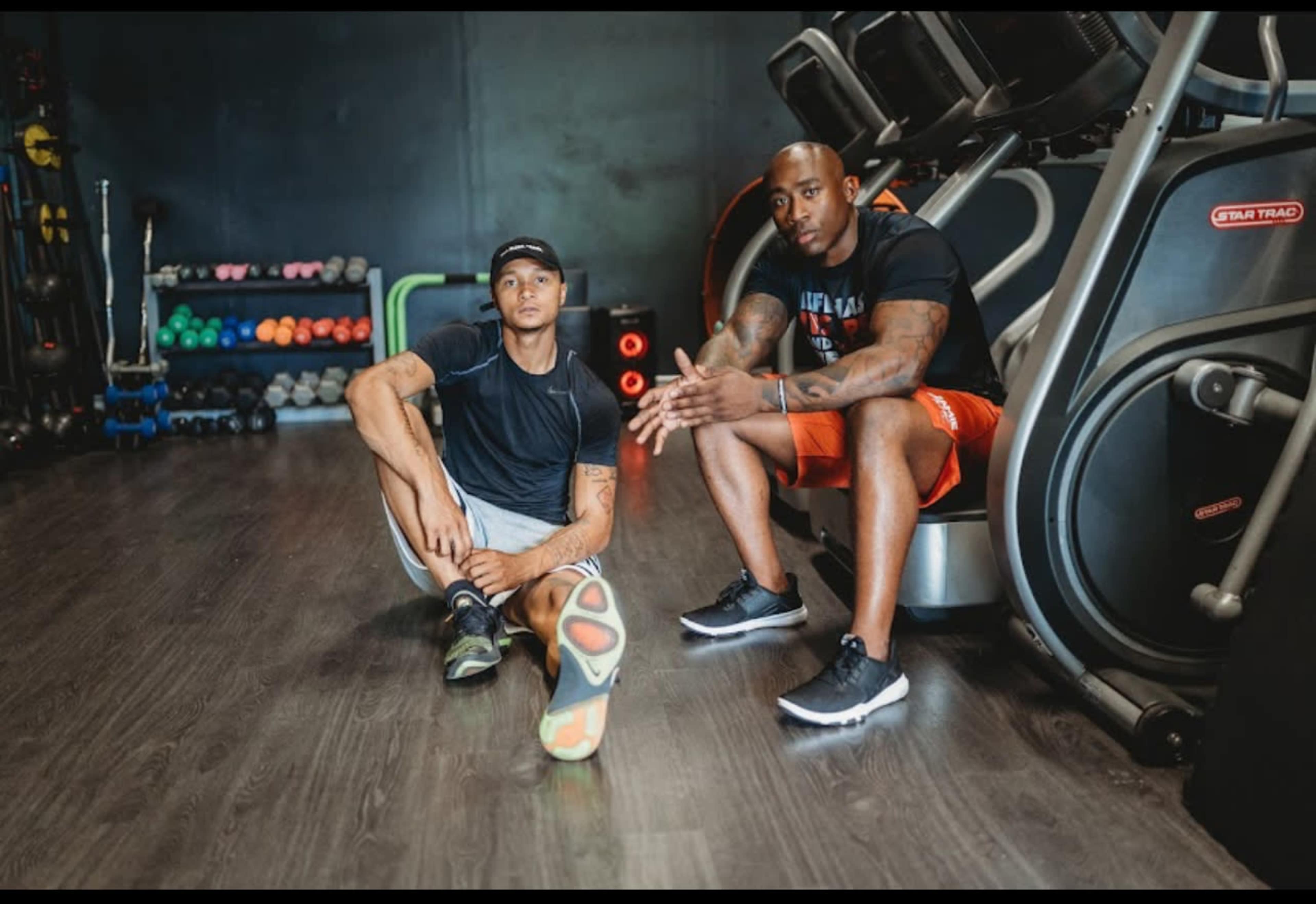 Two men are seated on a gym floor next to exercise equipment, one stretching his leg while the other sits with his hands clasped.