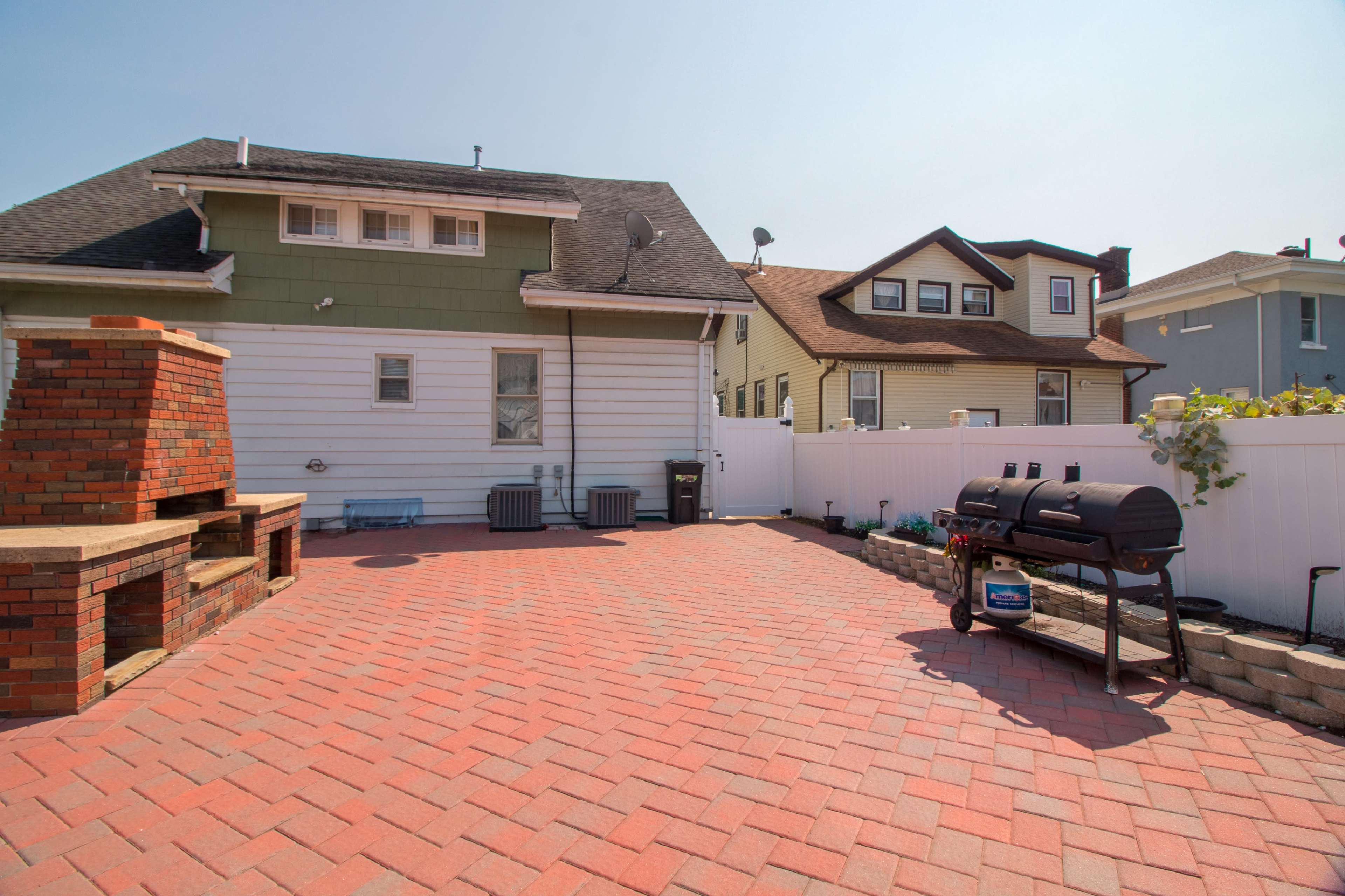 A brick-paved backyard with a barbecue grill, a smoker, and two houses in the background.