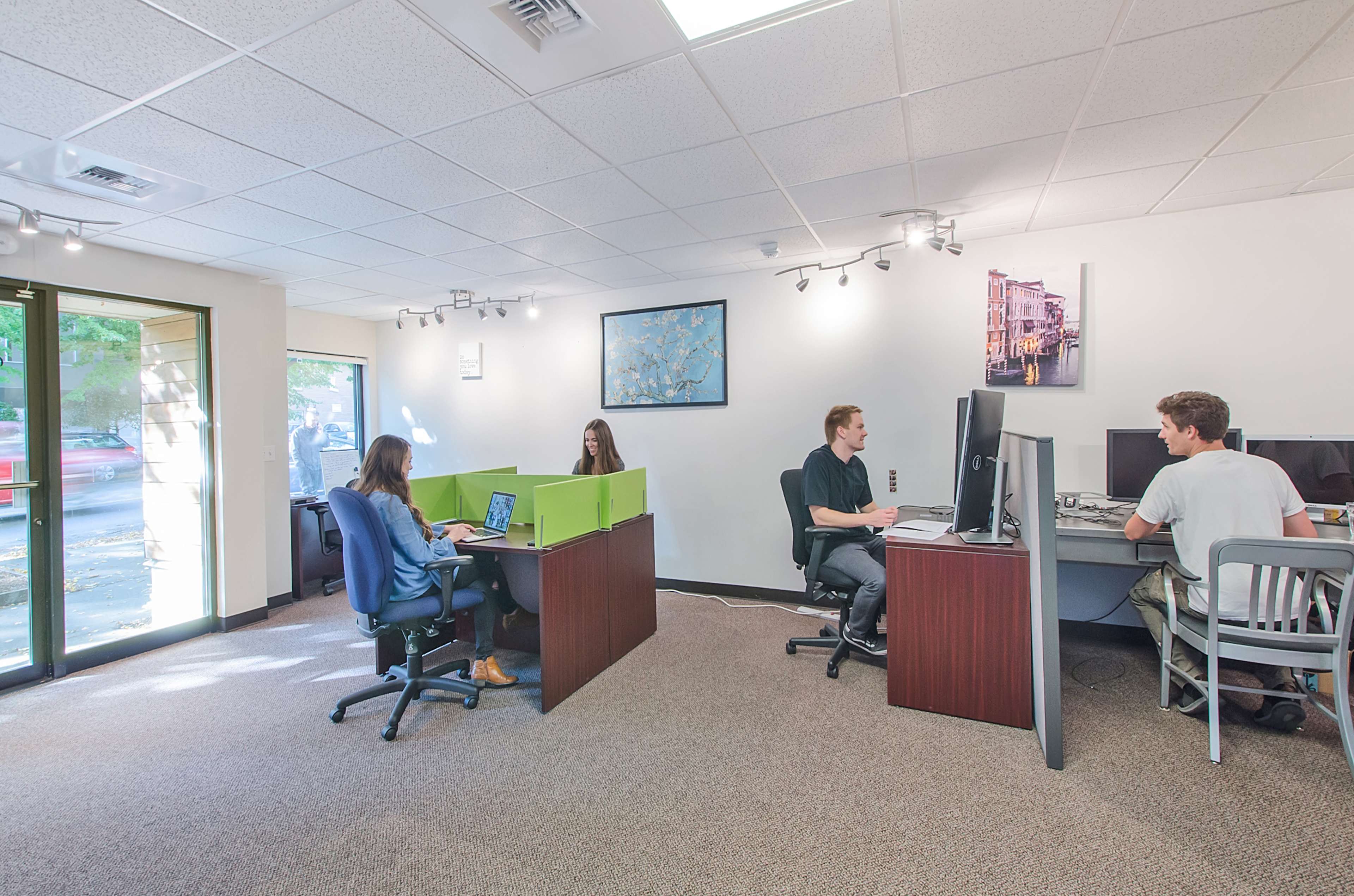 The image shows a modern office space with four individuals working at desks, separated by low partitions, and large windows providing natural light.