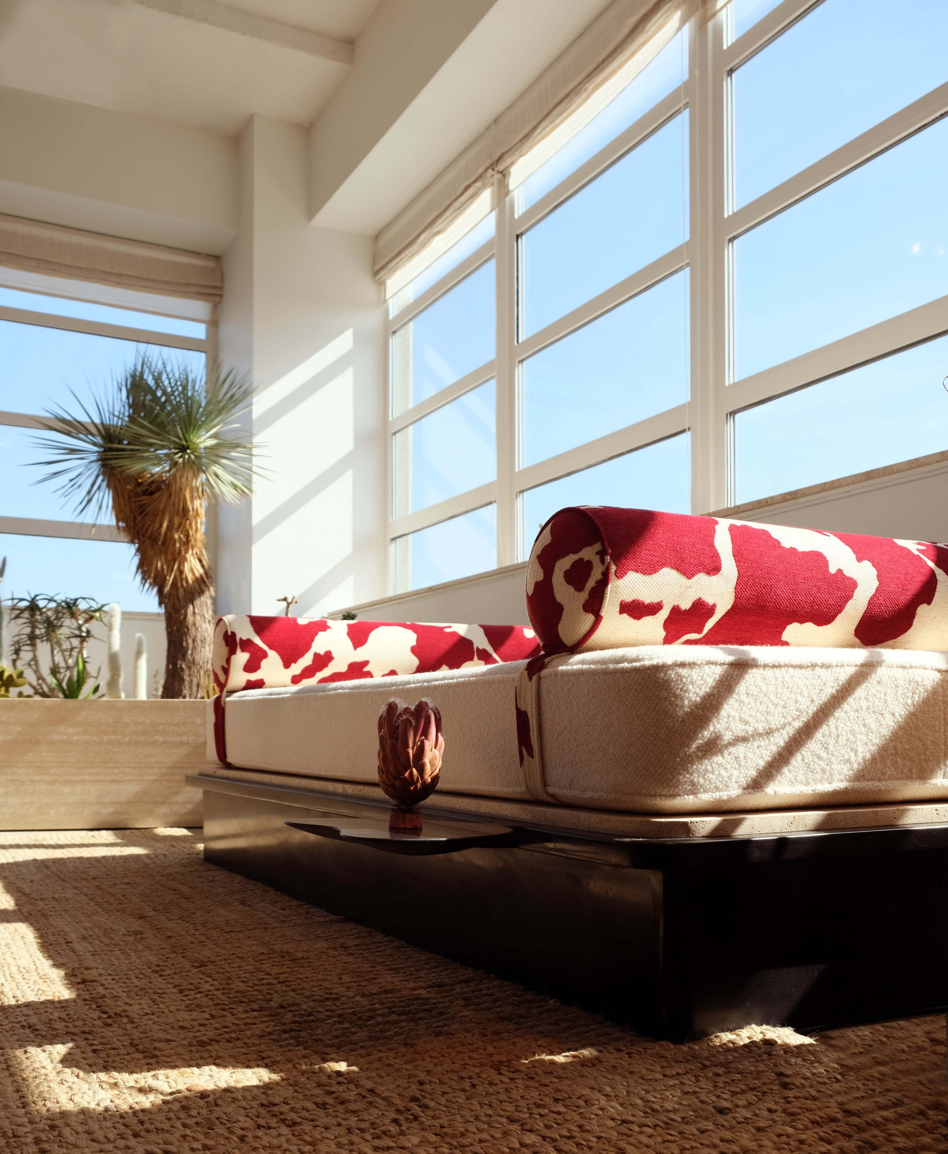 A modern living room features a light-colored sofa with bold red and white upholstery, surrounded by large windows that let in natural light and showcase a potted palm plant.