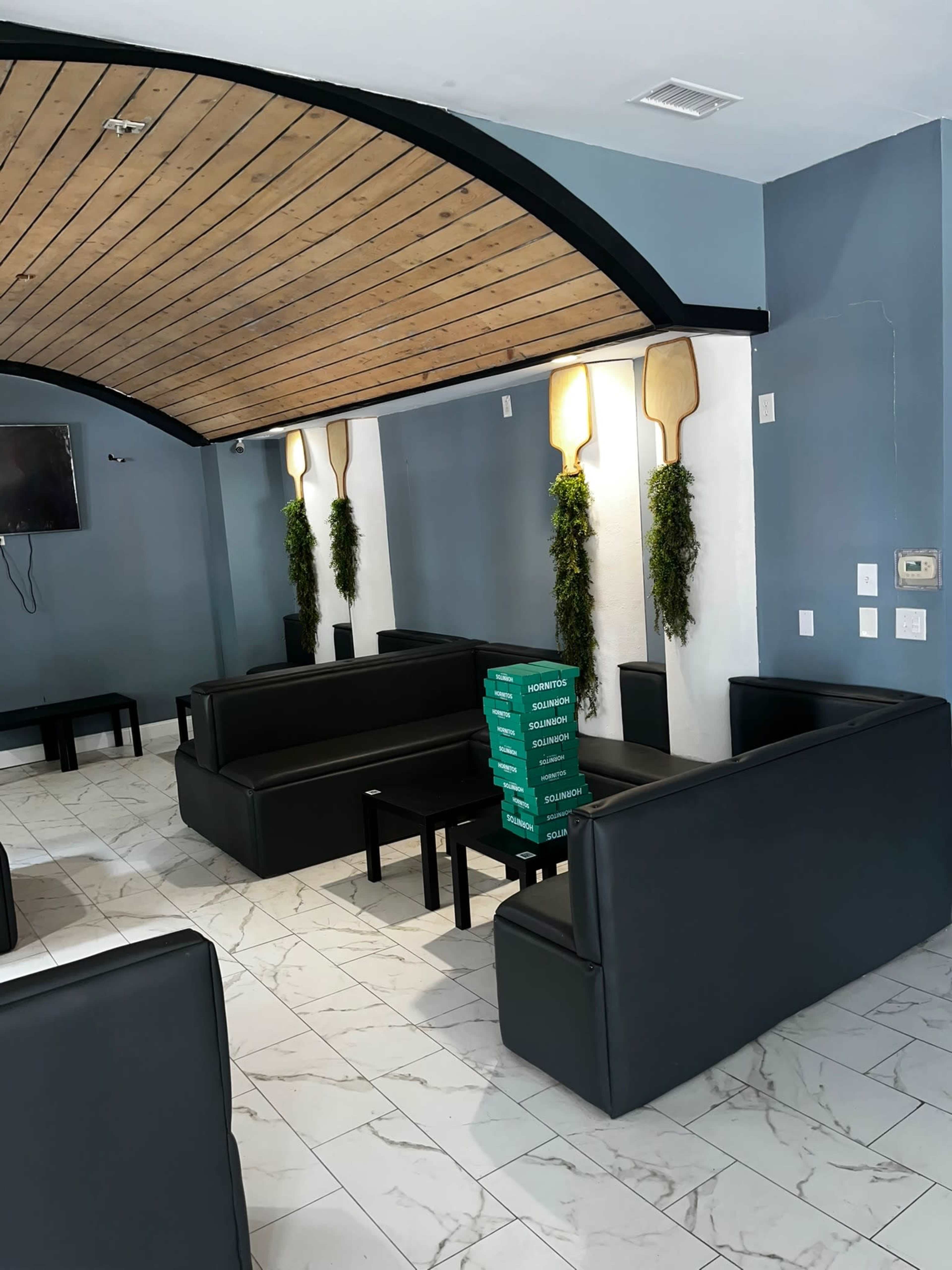 The image shows a modern lounge area with black leather sofas, small tables, and a decorative wooden ceiling, featuring green plants on the walls.
