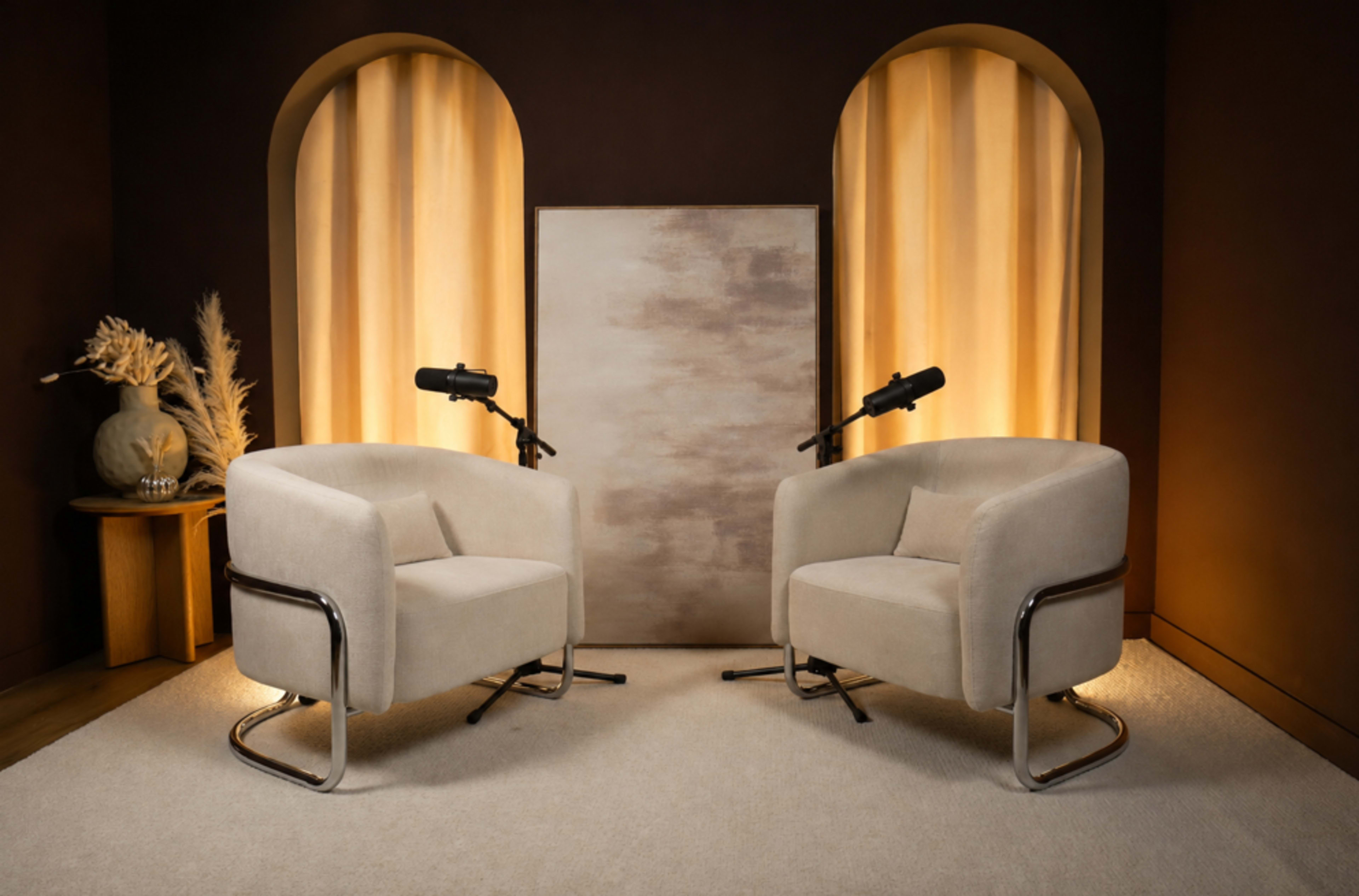 The image shows a modern, minimalistic interview setup with two beige armchairs facing each other, a neutral backdrop, and two microphones on stands positioned between them.