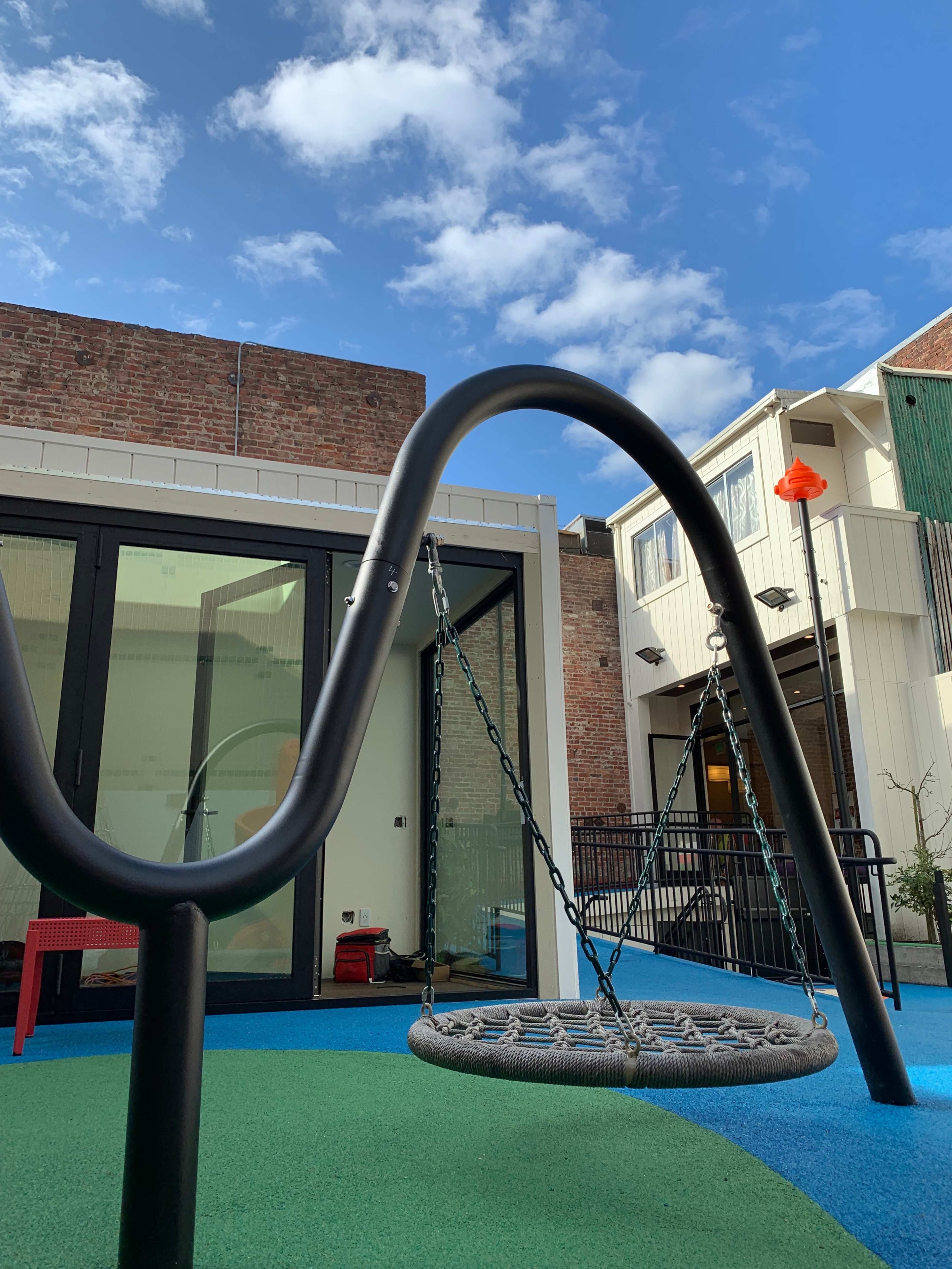 A black swing with a round seat is positioned in a colorful playground area near a building with large glass windows.