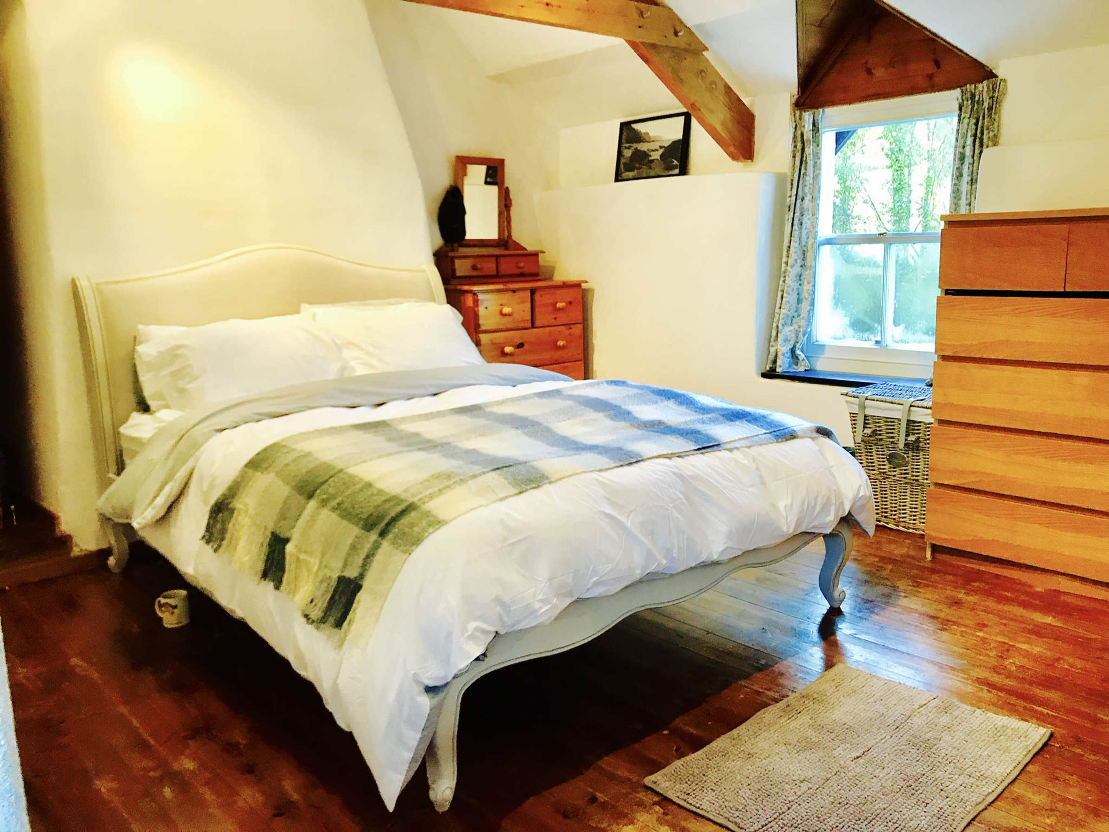 The image shows a cozy bedroom featuring a large bed with a plaid blanket, wooden floors, a window with curtains, and a chest of drawers.