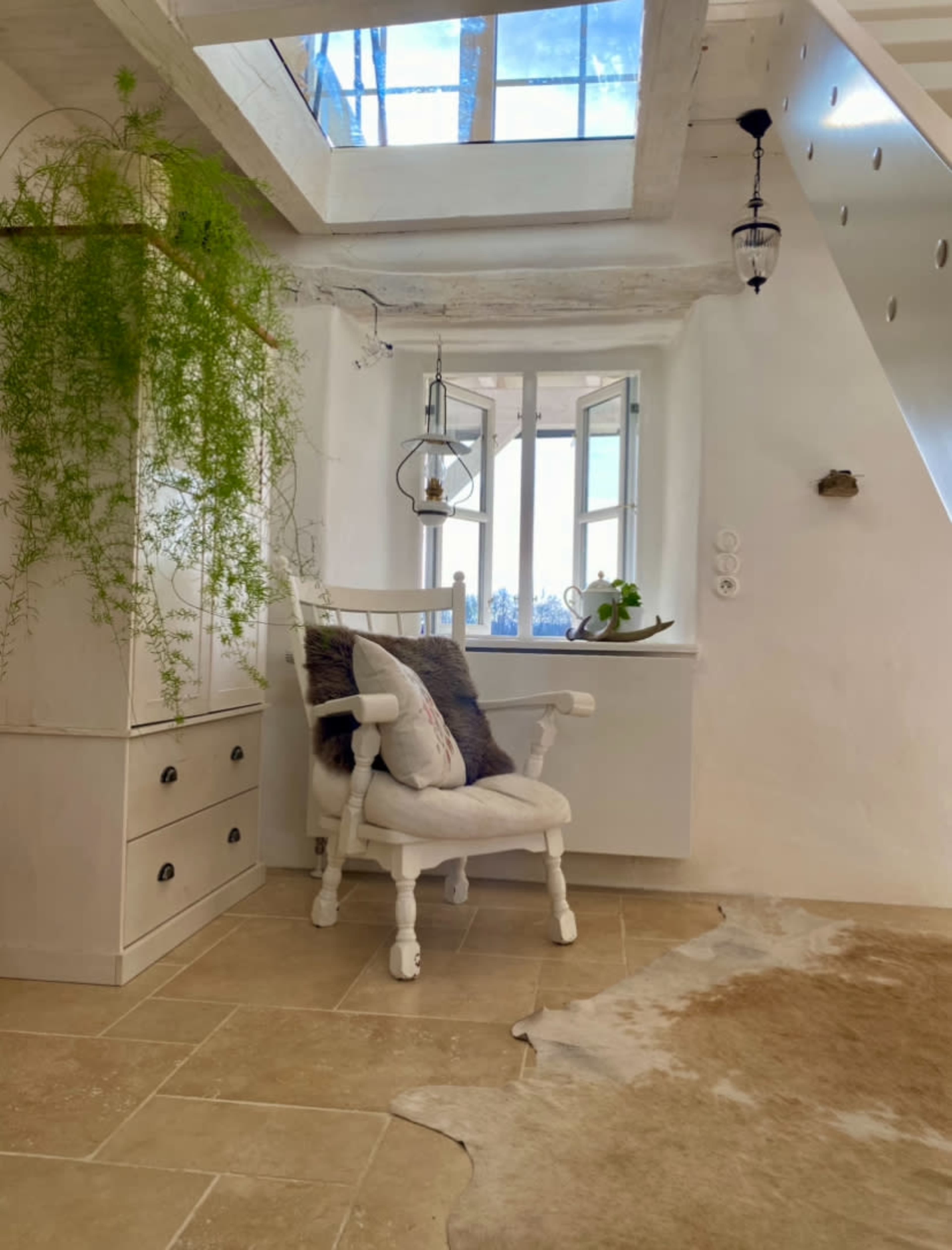 The image shows a bright interior space featuring a white chair next to a window, with potted plants and a soft rug on the tiled floor.