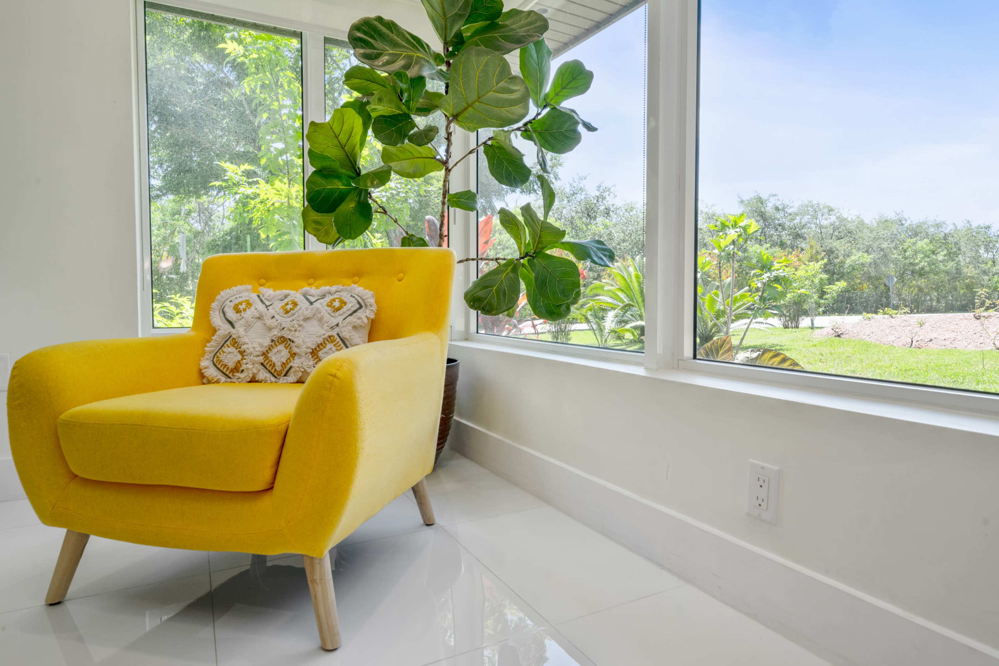 A yellow armchair with a patterned cushion is placed near a large window showcasing green plants and natural light.