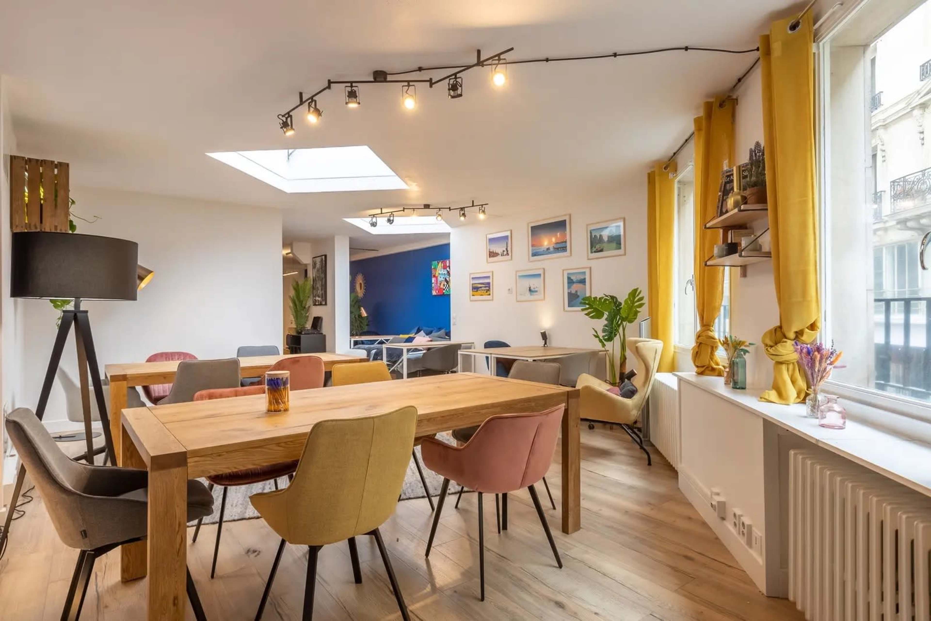 The scene shows a bright, open multi-purpose room featuring a wooden dining table surrounded by colorful chairs, large windows with yellow curtains, and various wall decorations including photographs.