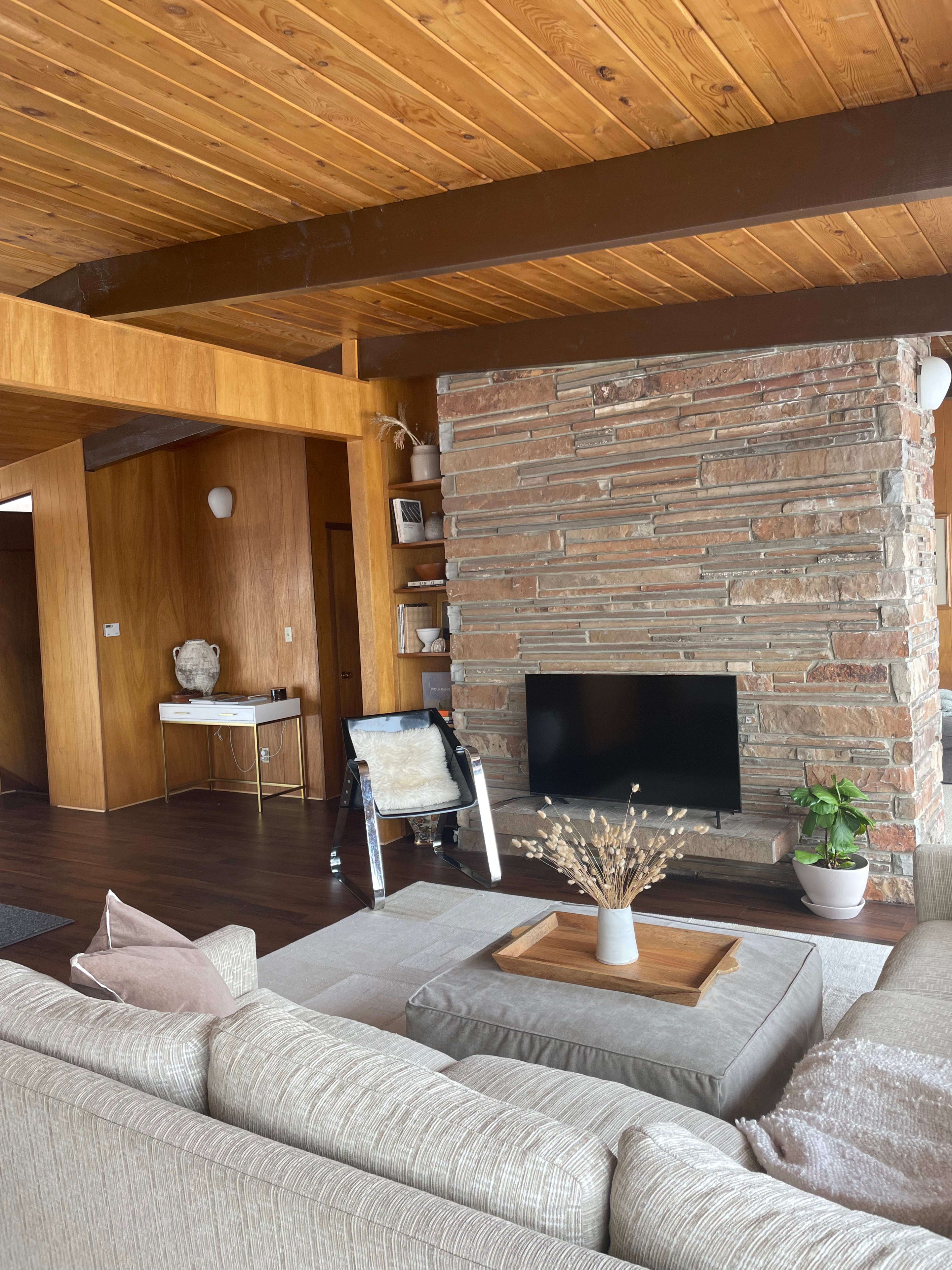 The living room features a stone fireplace, a large beige sofa, and wooden beams on the ceiling, with a television mounted on a stone wall.