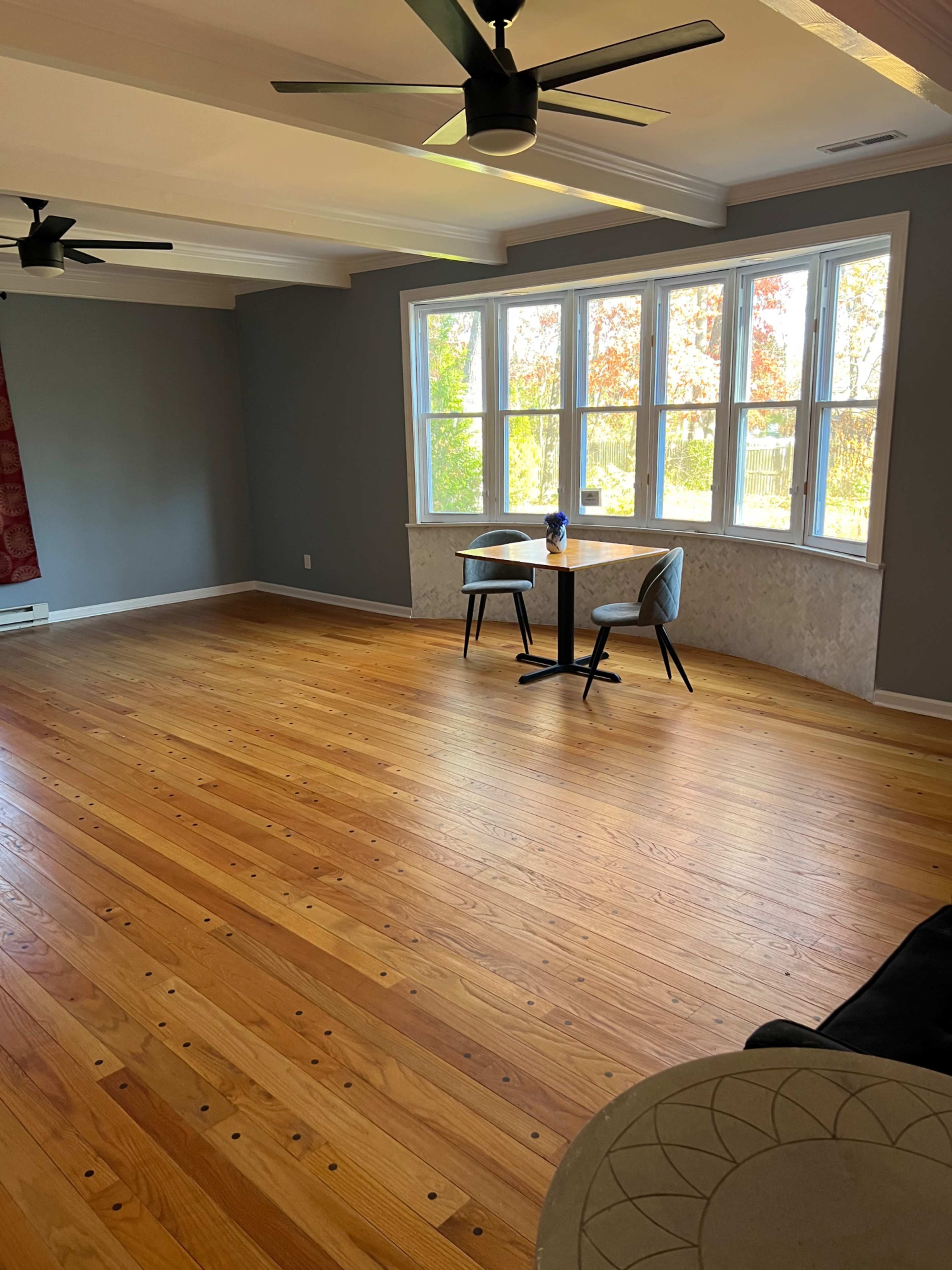 A room features polished wooden floors, two ceiling fans, large windows letting in natural light, and a small dining table with two chairs.