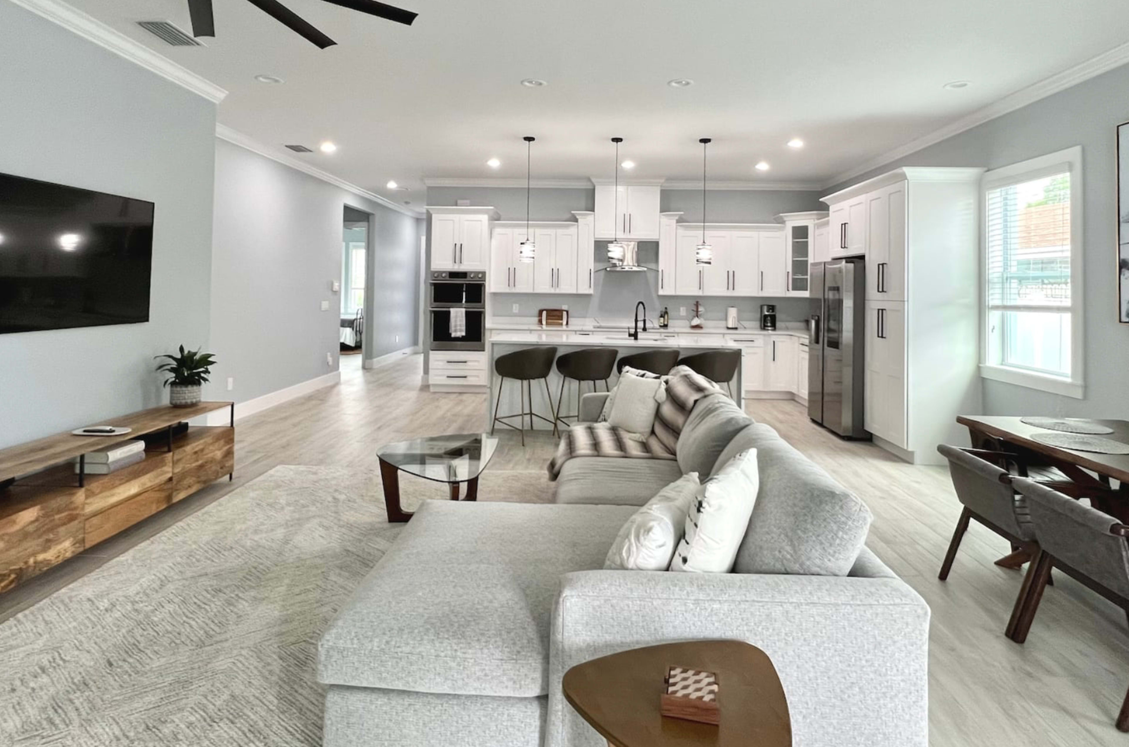 A modern, open-concept living space with a gray and white color scheme, featuring a sectional sofa, a kitchen with an island, and a dining area.