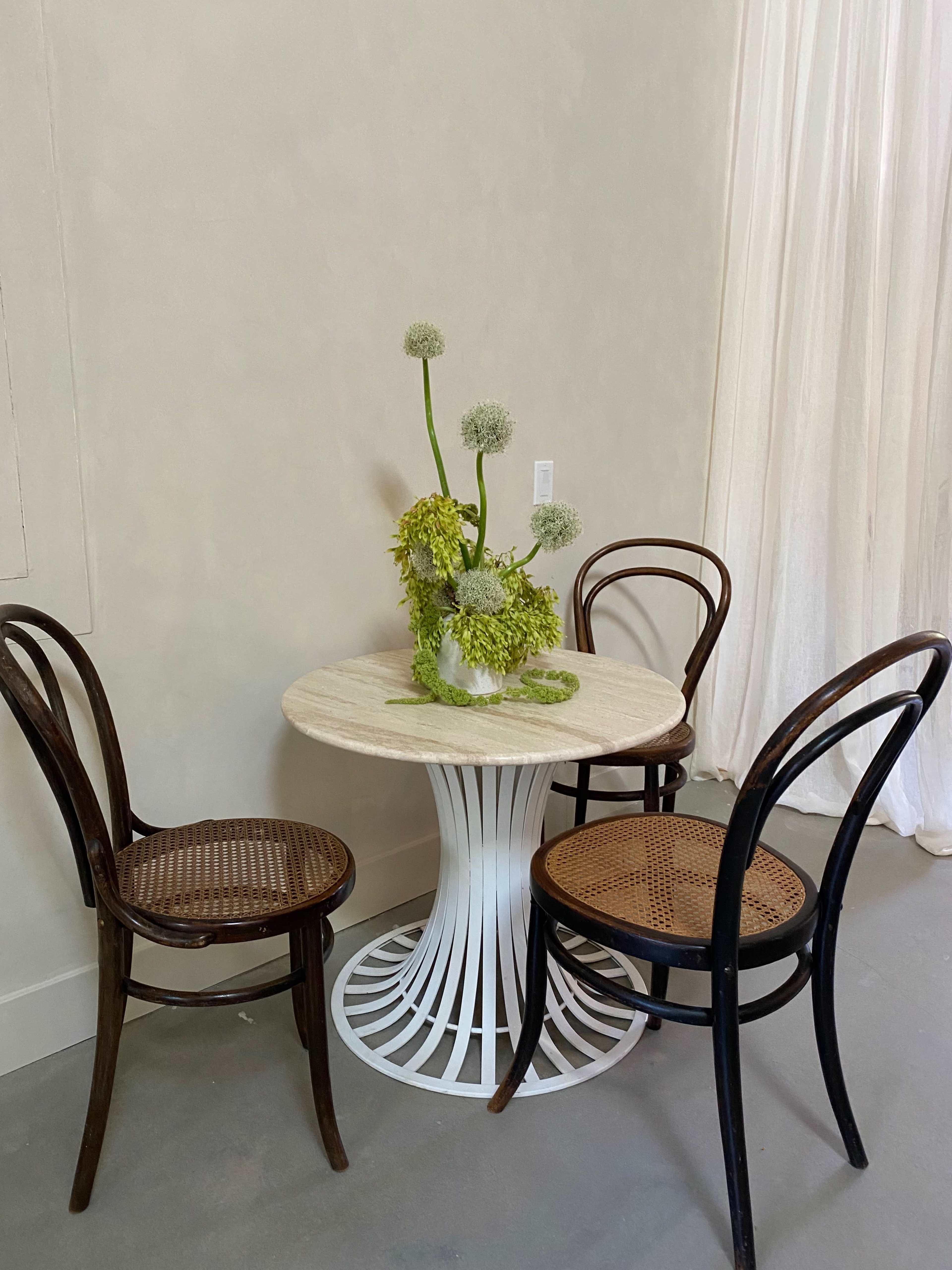 A round table with a white pedestal base is surrounded by two wooden chairs, with a floral arrangement in a vase on the tabletop.